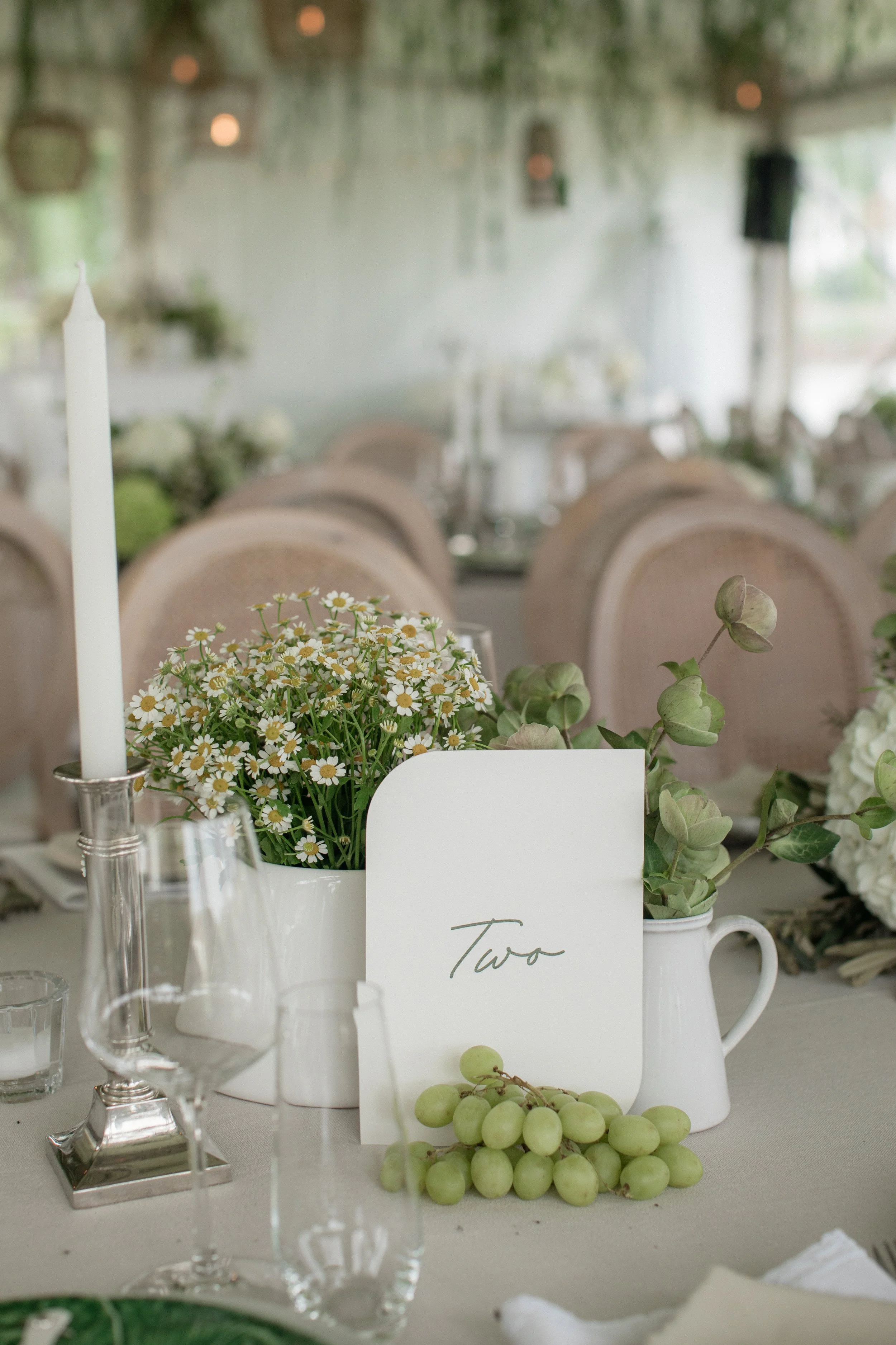 A table decorated for a wedding or celebration, with a white pitcher holding a bouquet of small white daisy flowers, a cluster of green grapes, a white card with the word "Two" written on it, and glassware. The background shows a blurred indoor setti