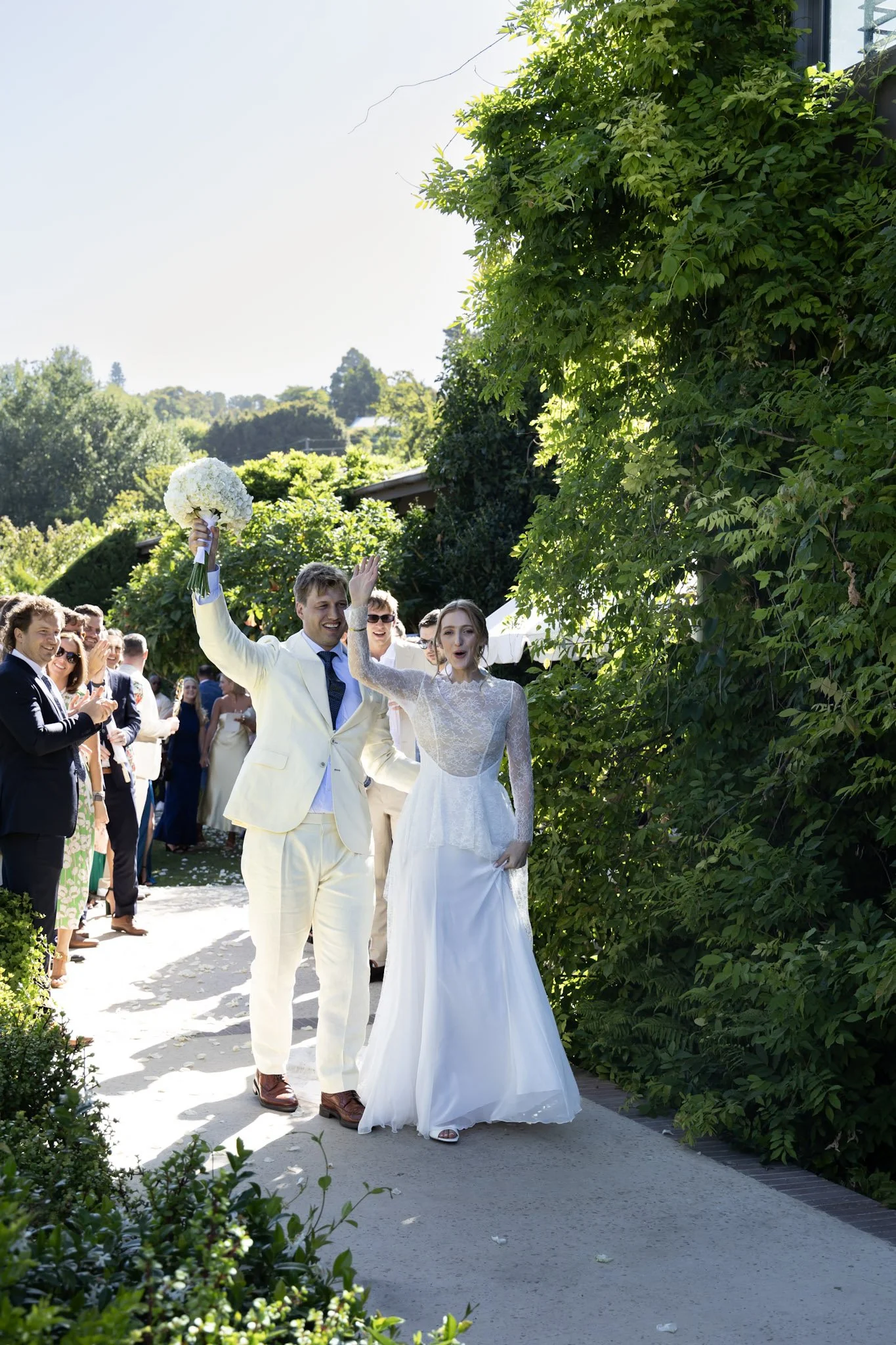 A wedding celebration taking place outdoors in bright sunlight, with a bride and groom walking down an aisle, surrounded by guests, lush green foliage, and trees in the background.