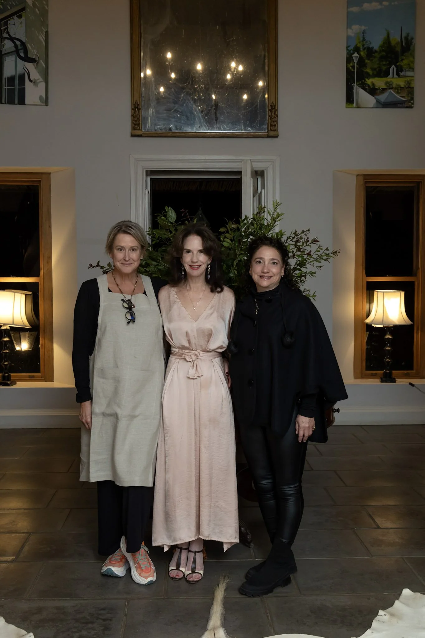 Three women standing indoors in front of a wall decorated with paintings and a mirror, with furniture including lamps on either side. They are smiling and posing for the photo, with a dog's tail visible at the bottom of the frame.