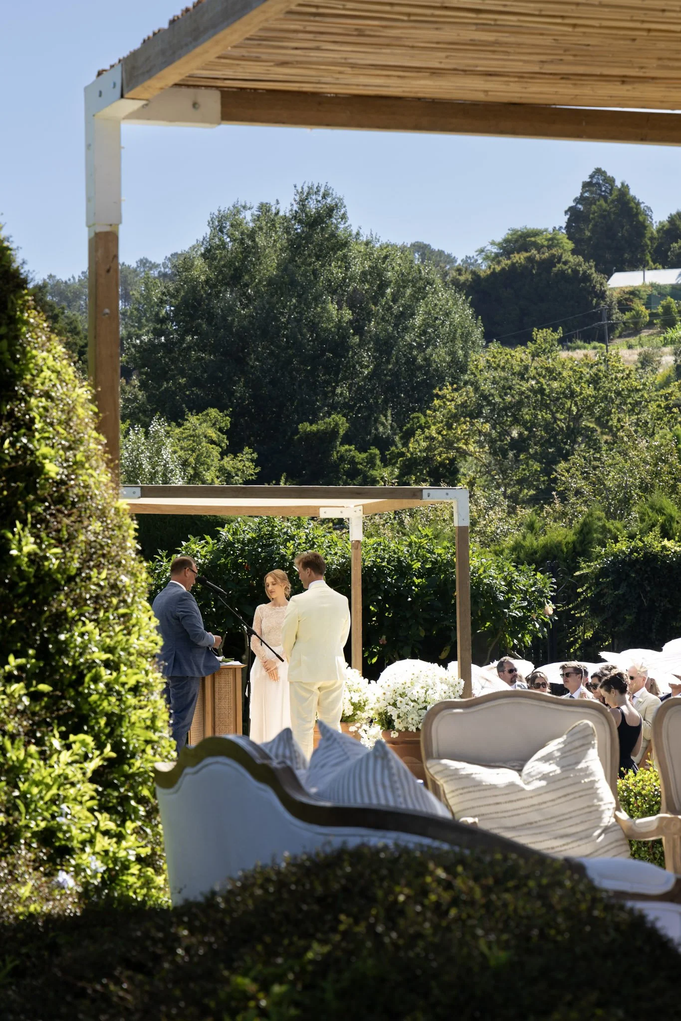 A wedding ceremony outdoors with a bride and groom facing each other, officiant reading, guests seated, surrounded by greenery and white flowers, under a wooden pergola on a sunny day.