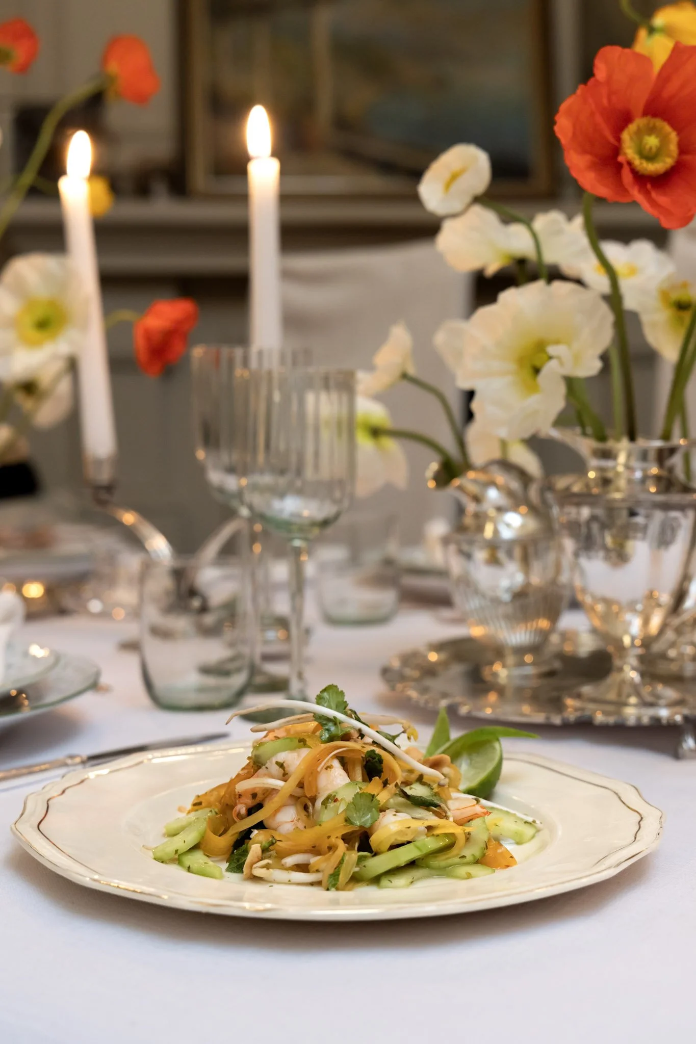 Plate of Asian stir-fried noodles with vegetables and shrimp on a white table, with a formal dining setup and floral centerpieces with candles and flowers in the background.