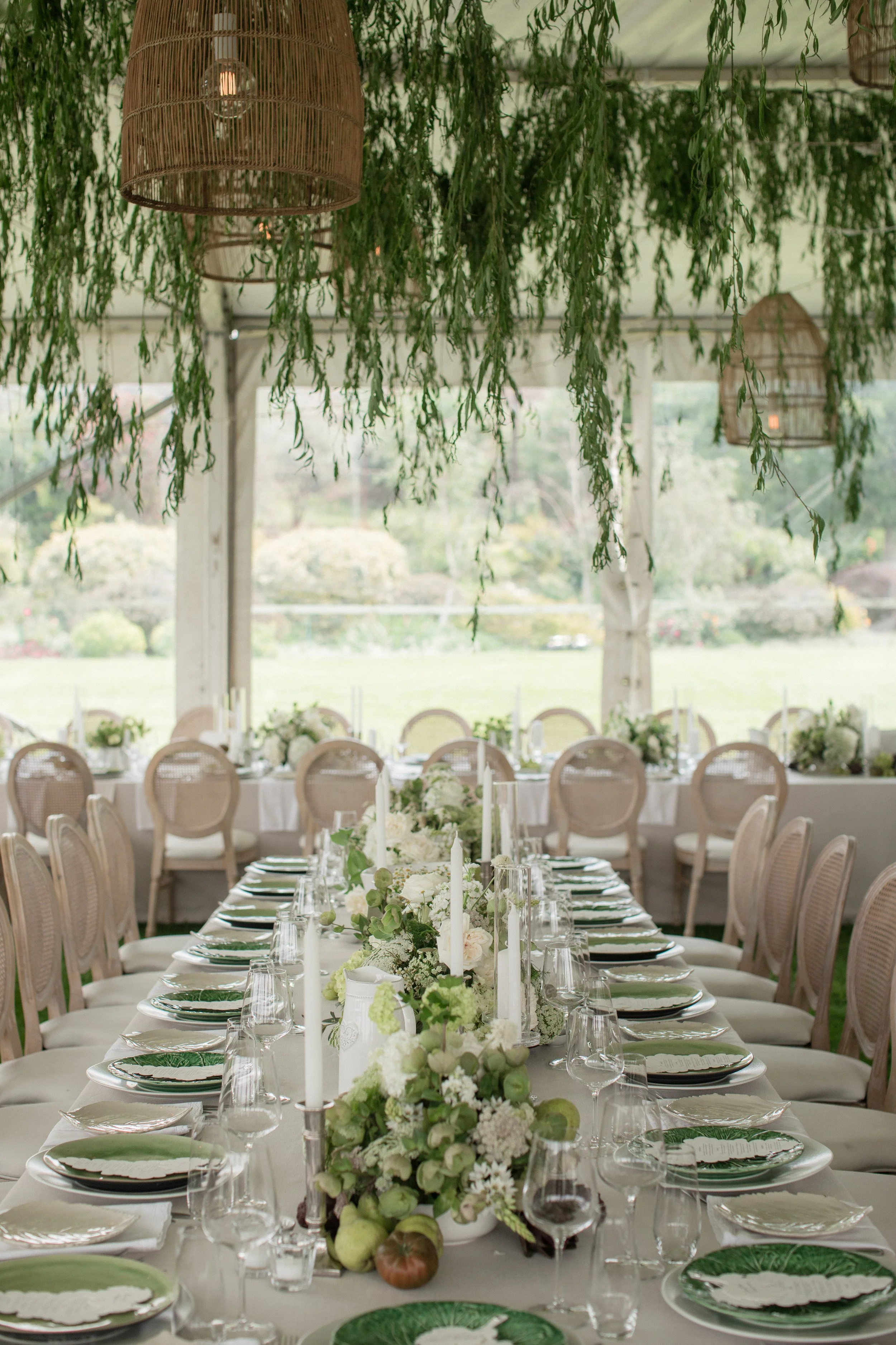 Elegant wedding reception table setup with white linens, floral centerpieces, candles, and green tableware, under hanging greenery and wicker lanterns.
