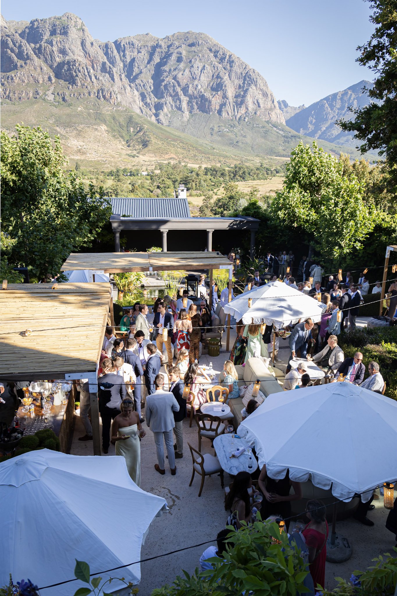 A lively outdoor gathering or celebration with many people dressed in formal attire, under white umbrellas, with a scenic mountain backdrop and lush greenery.
