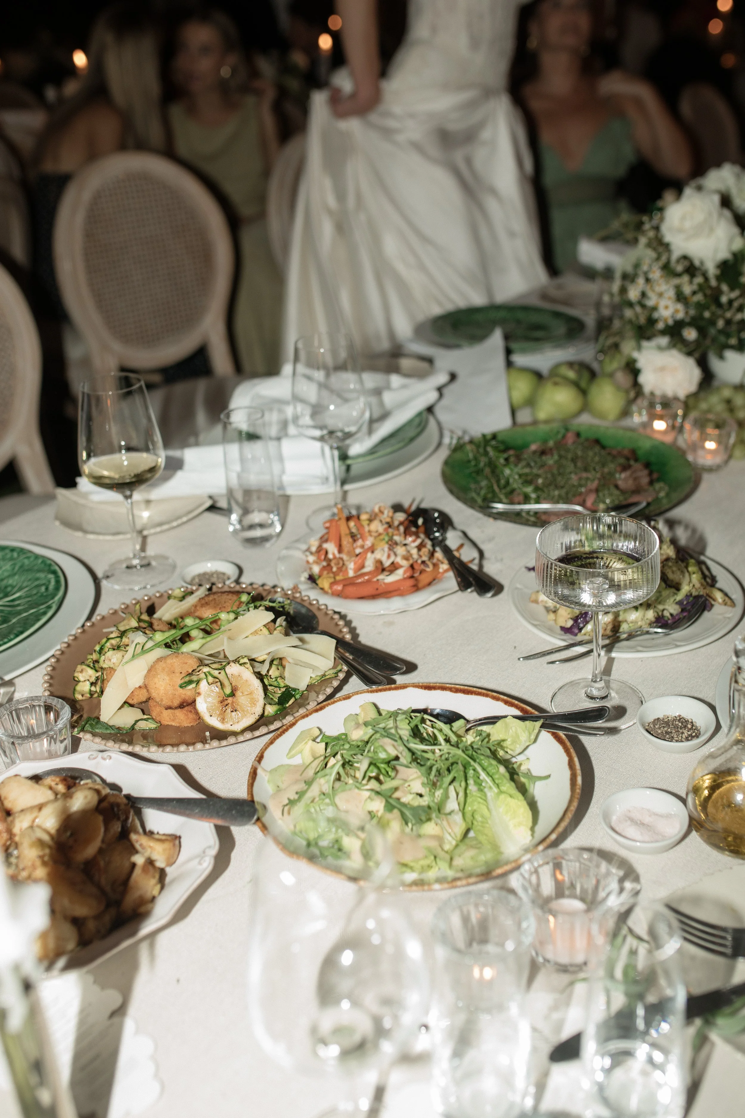 A dinner table set with various dishes including salads, roasted vegetables, and a plate with lemon slices, surrounded by glasses of wine and water, with people in formal attire in the background at a banquet or celebration.