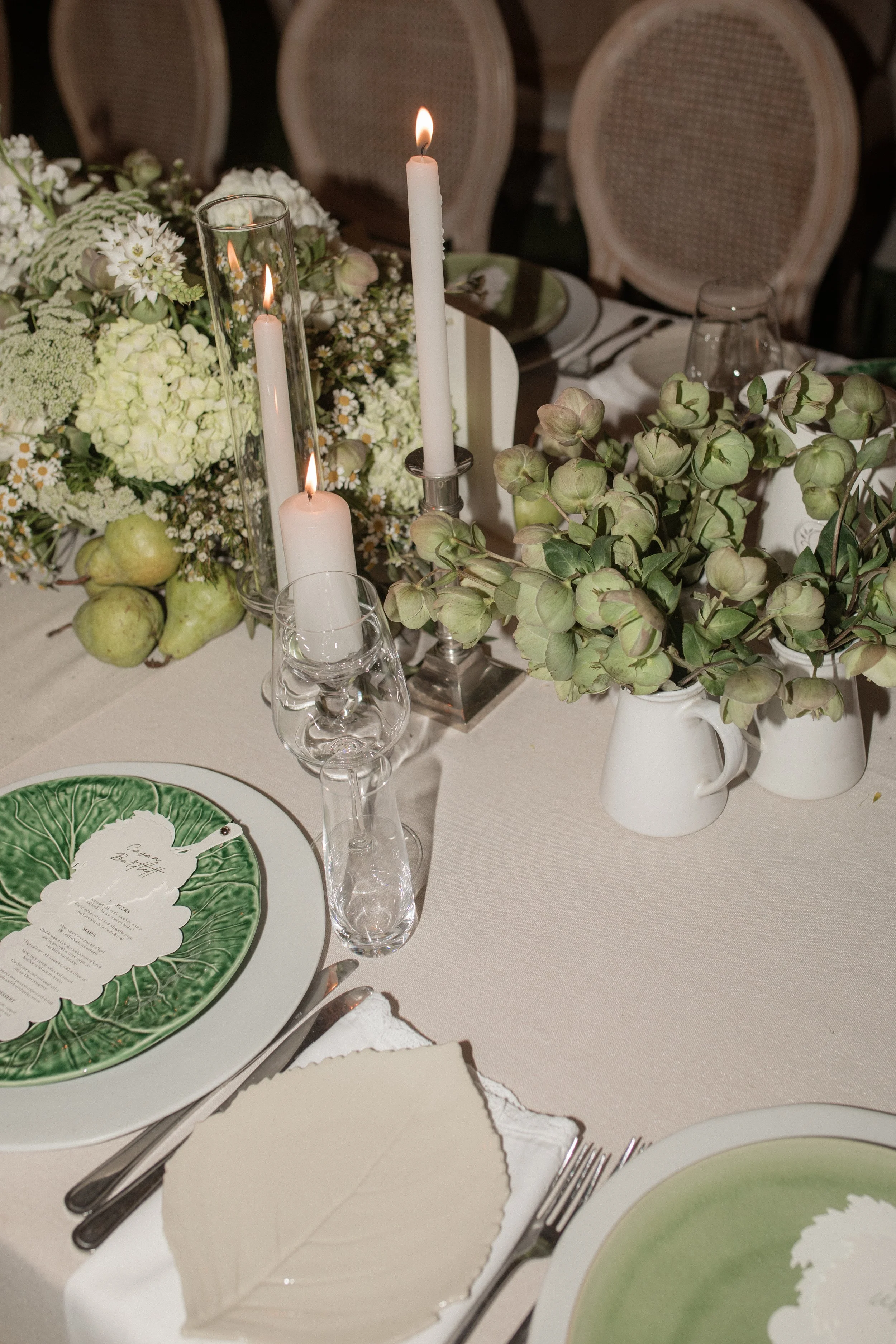 A table setting with white candles, a bouquet of white and green flowers, green pears, and elegant dishware. The table is decorated for a special occasion, with a menu card on a green and white plate.