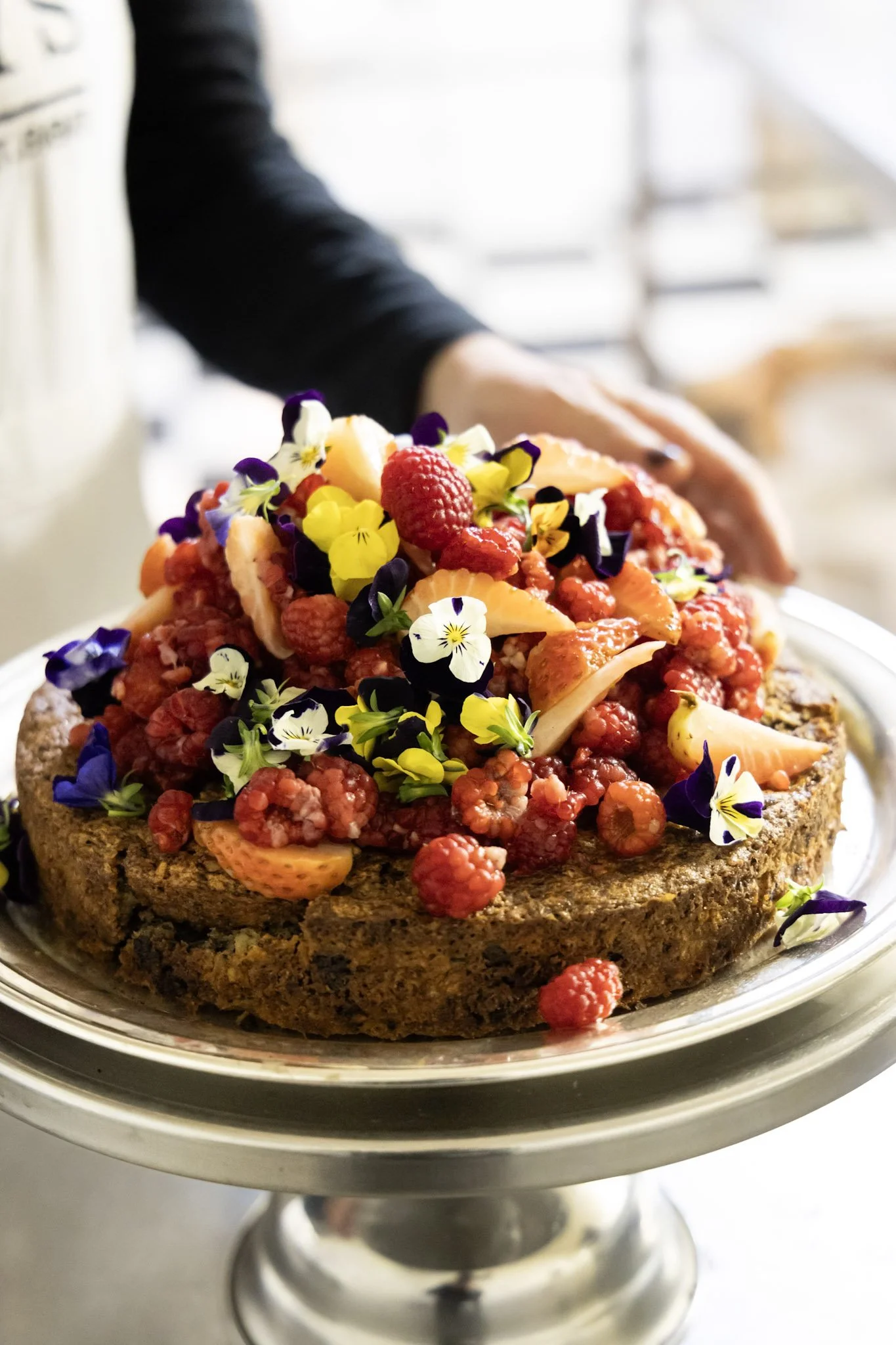 A cake topped with mixed berries, edible flowers, and fruit slices in a decorative presentation on a silver cake stand.