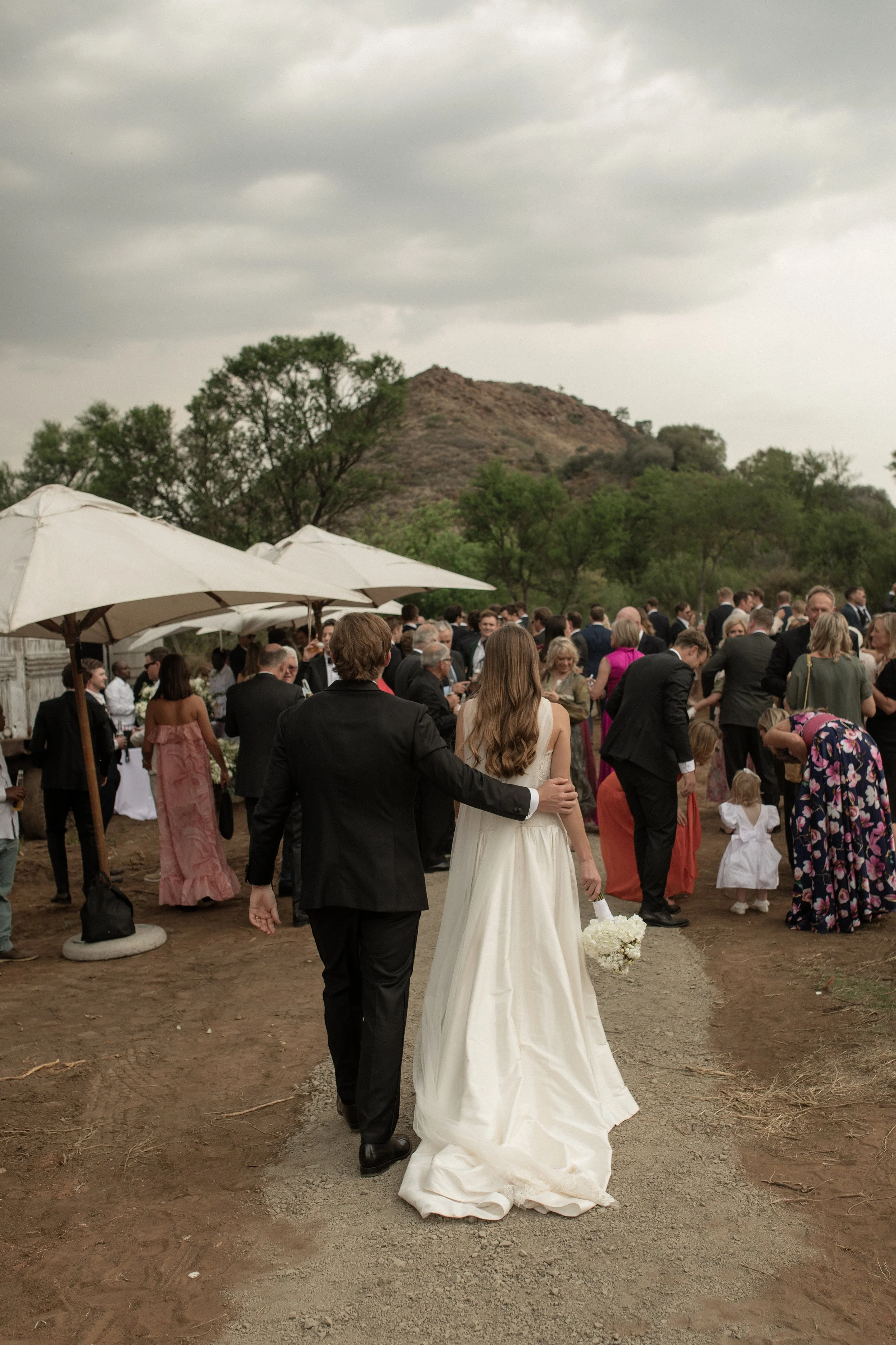 A wedding reception outdoors with a bride in a white gown and a groom in a black tuxedo walking away from the camera. The scene includes many guests mingling, some of whom are dressed in formal attire. There are large white umbrellas providing shade,