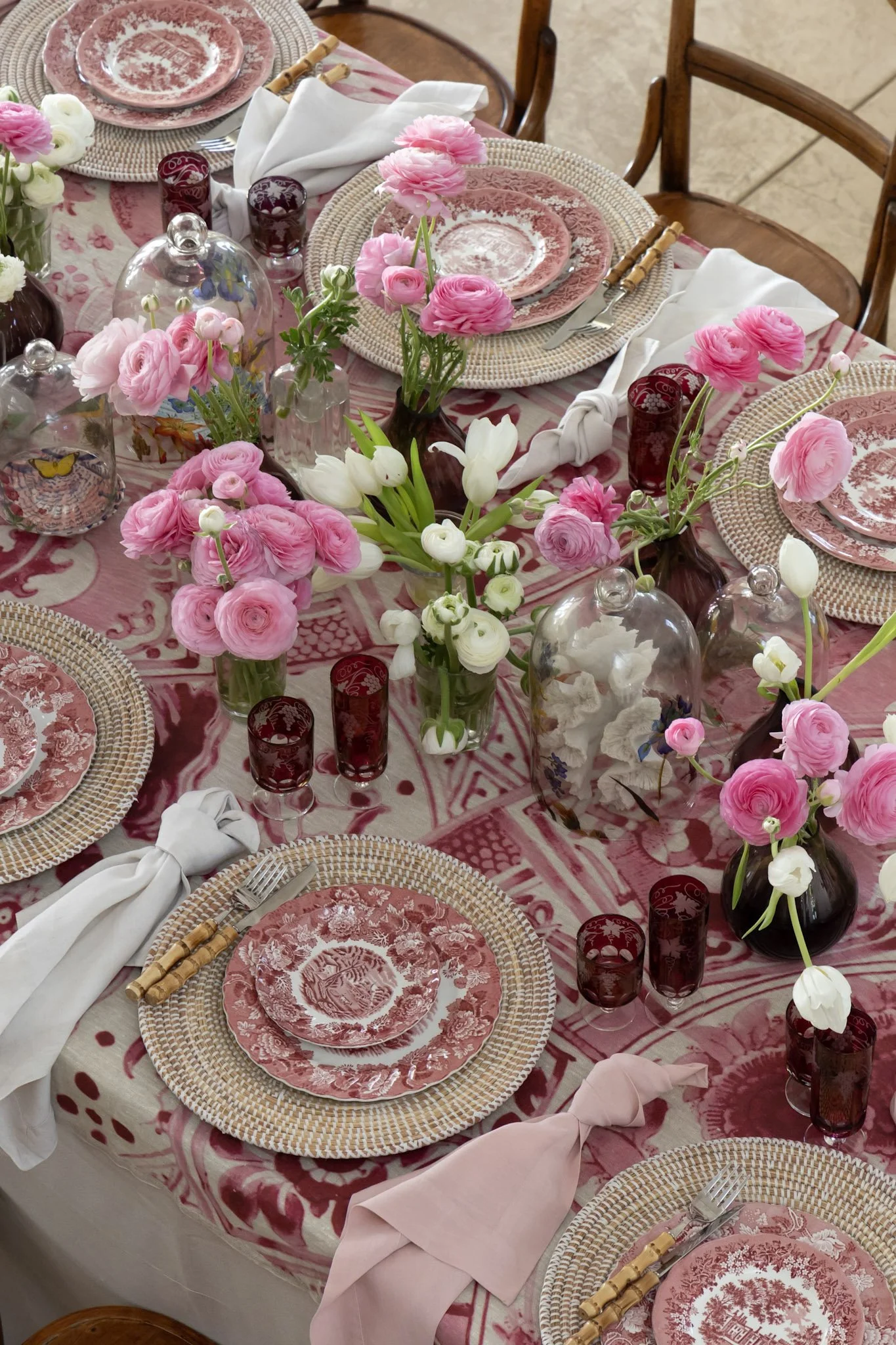A table set for a meal with pink and white flowers, pink patterned plates, woven placemats, white napkins, and red glasses, in a pink and white themed decor.