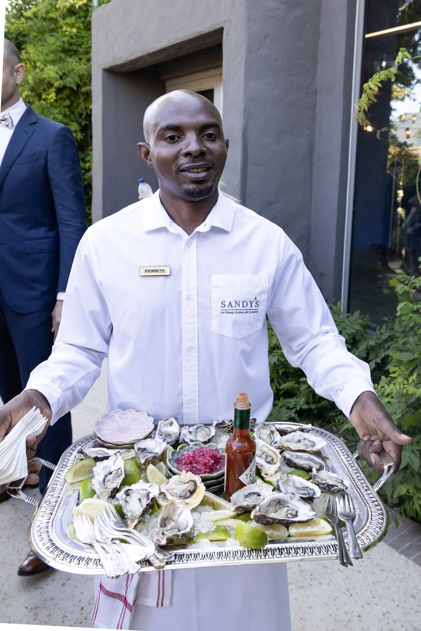 Man in white shirt with a name tag serving oysters on a silver tray with lemon wedges and hot sauce, outdoors with greenery and a gray building in the background.