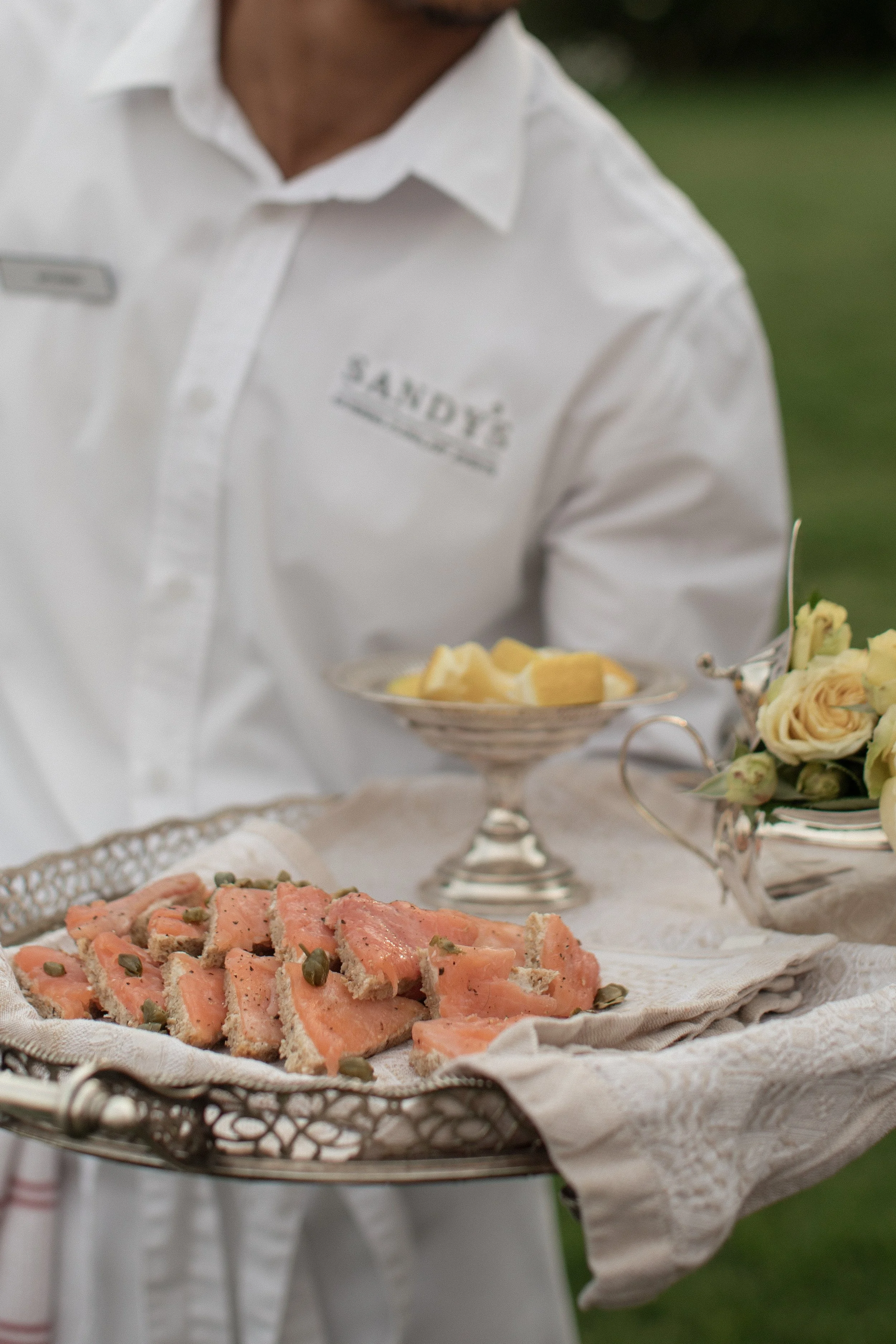 A person dressed in a white shirt with a name tag that reads 'SANDY' is holding a silver tray with slices of smoked salmon garnished with capers. There are also flowers and other food items on the table.