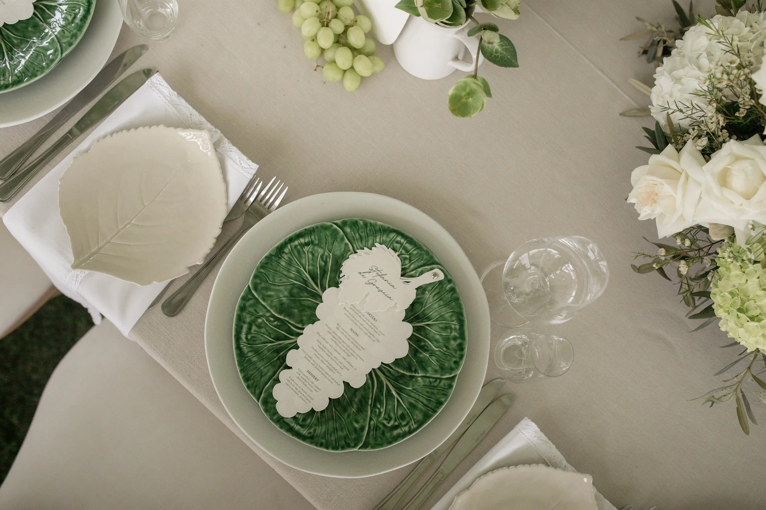 A table setting with green leaf-patterned plates, a menu card, silverware, wine glasses, and floral arrangements with white flowers and greenery.