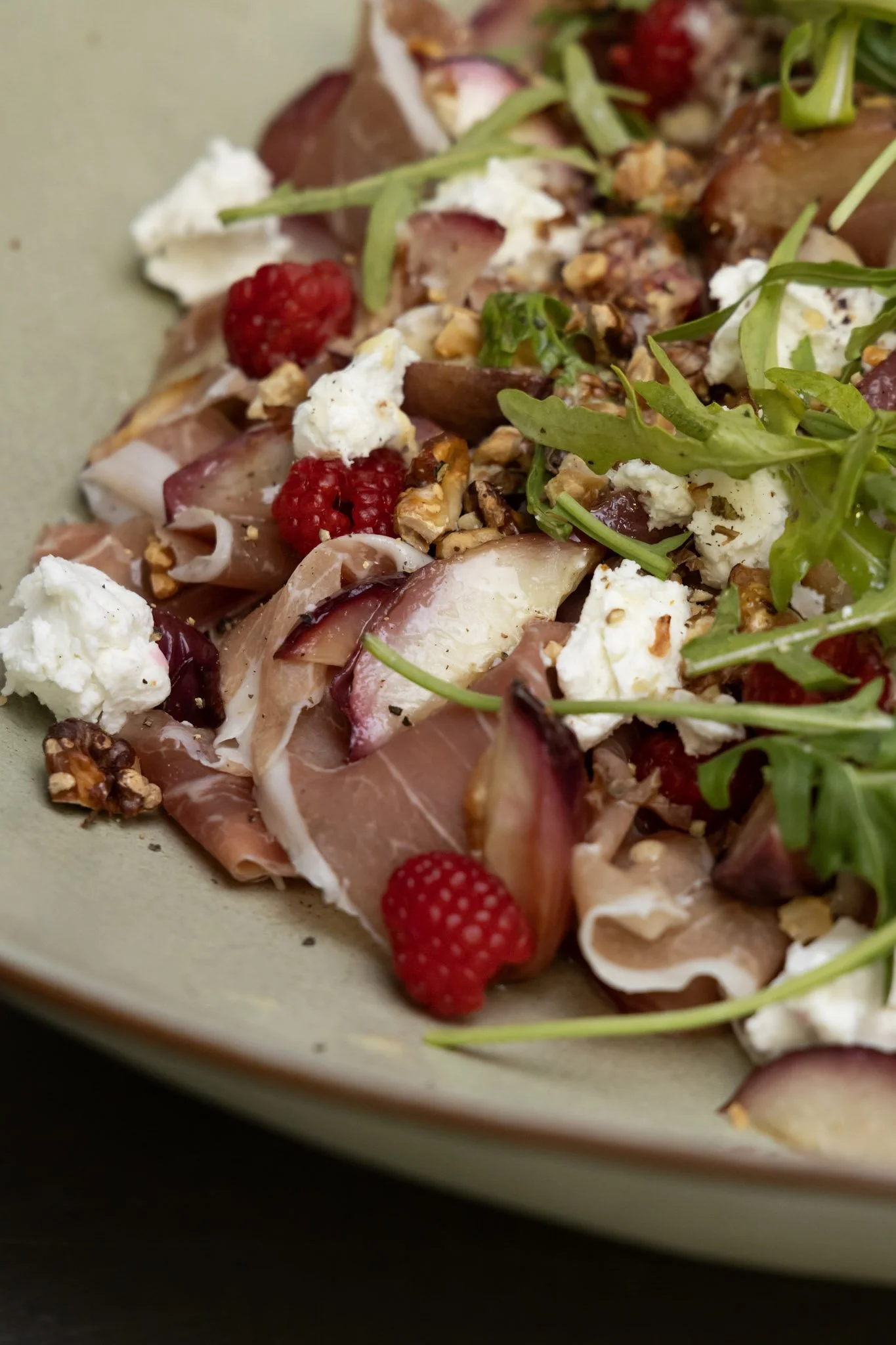 Close-up of a gourmet dish with raspberries, cheese, green garnishes, nuts, and thin slices of meat on an off-white plate.