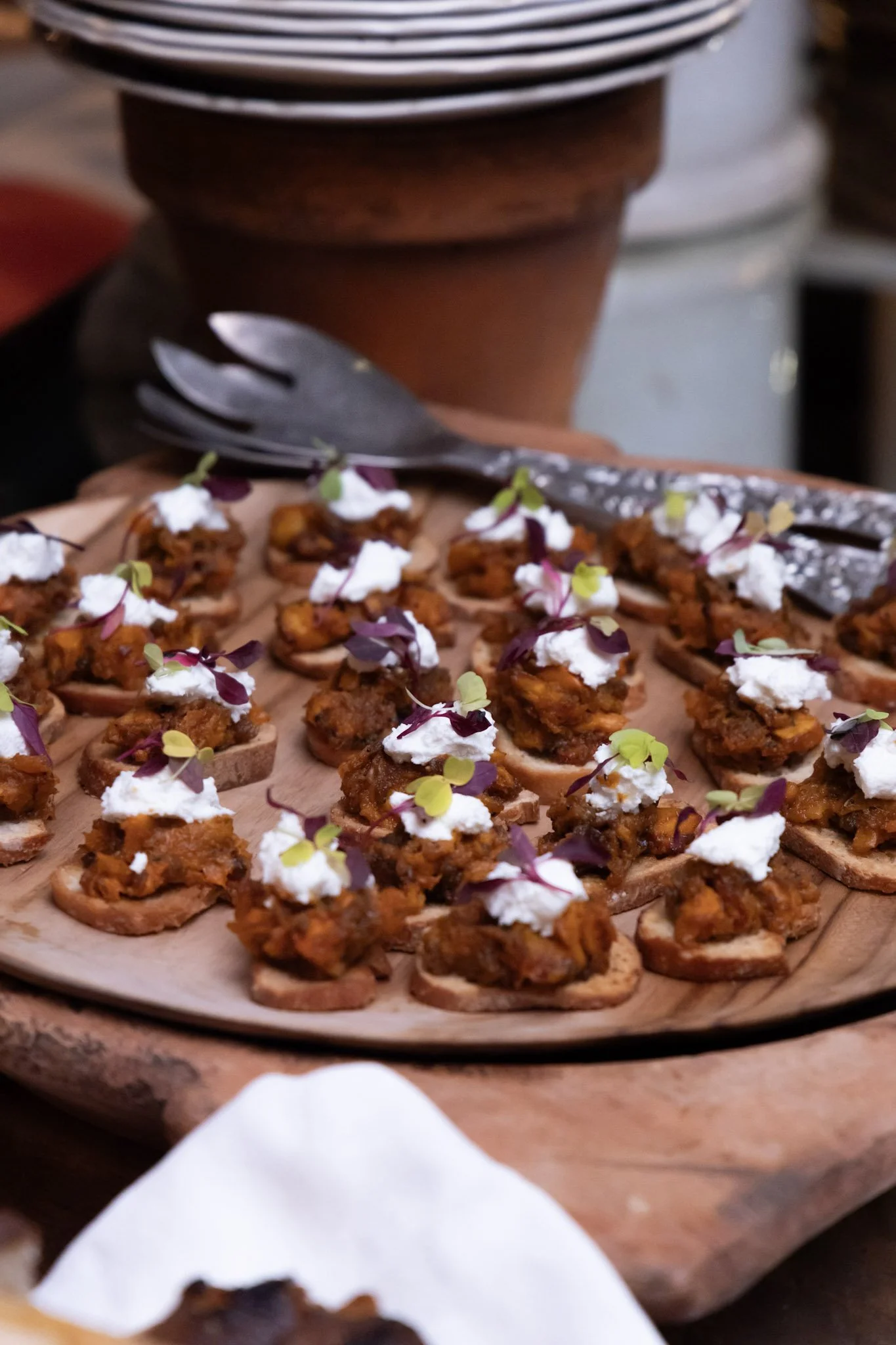 Close-up of small appetizers with a dollop of white sauce and microgreens on a wooden serving platter. In the background, a stack of dishes and serving utensils are visible.