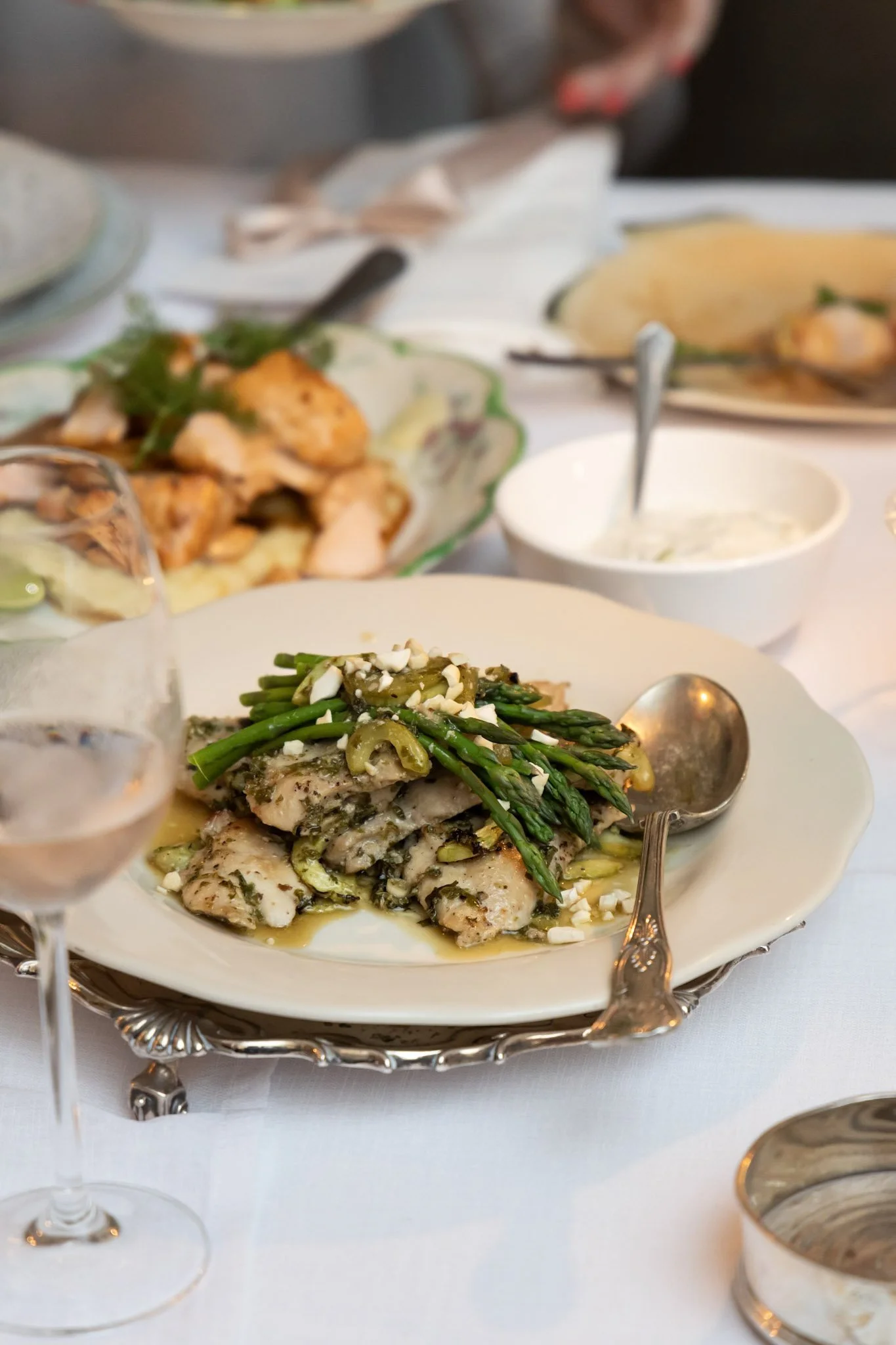 A plated dish with fish topped with green beans and chopped nuts on a silver tray. In the background, several other dishes and a glass of wine are on a white tablecloth.