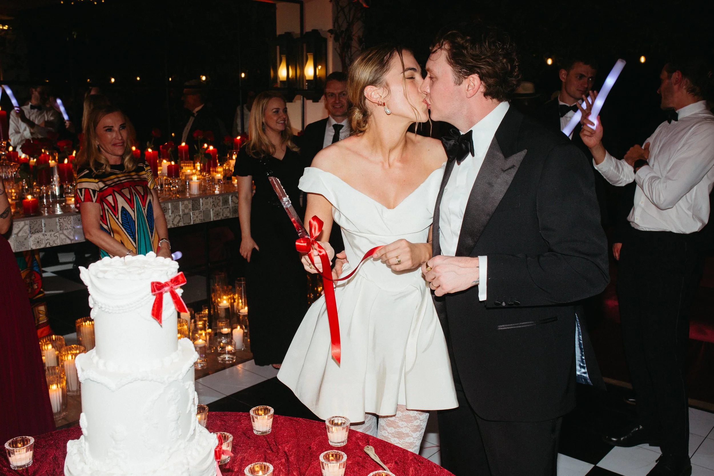 A wedding reception with a bride and groom sharing a kiss near a wedding cake decorated with red ribbons. Guests in formal attire are smiling and watching, with candles and candles in the background.