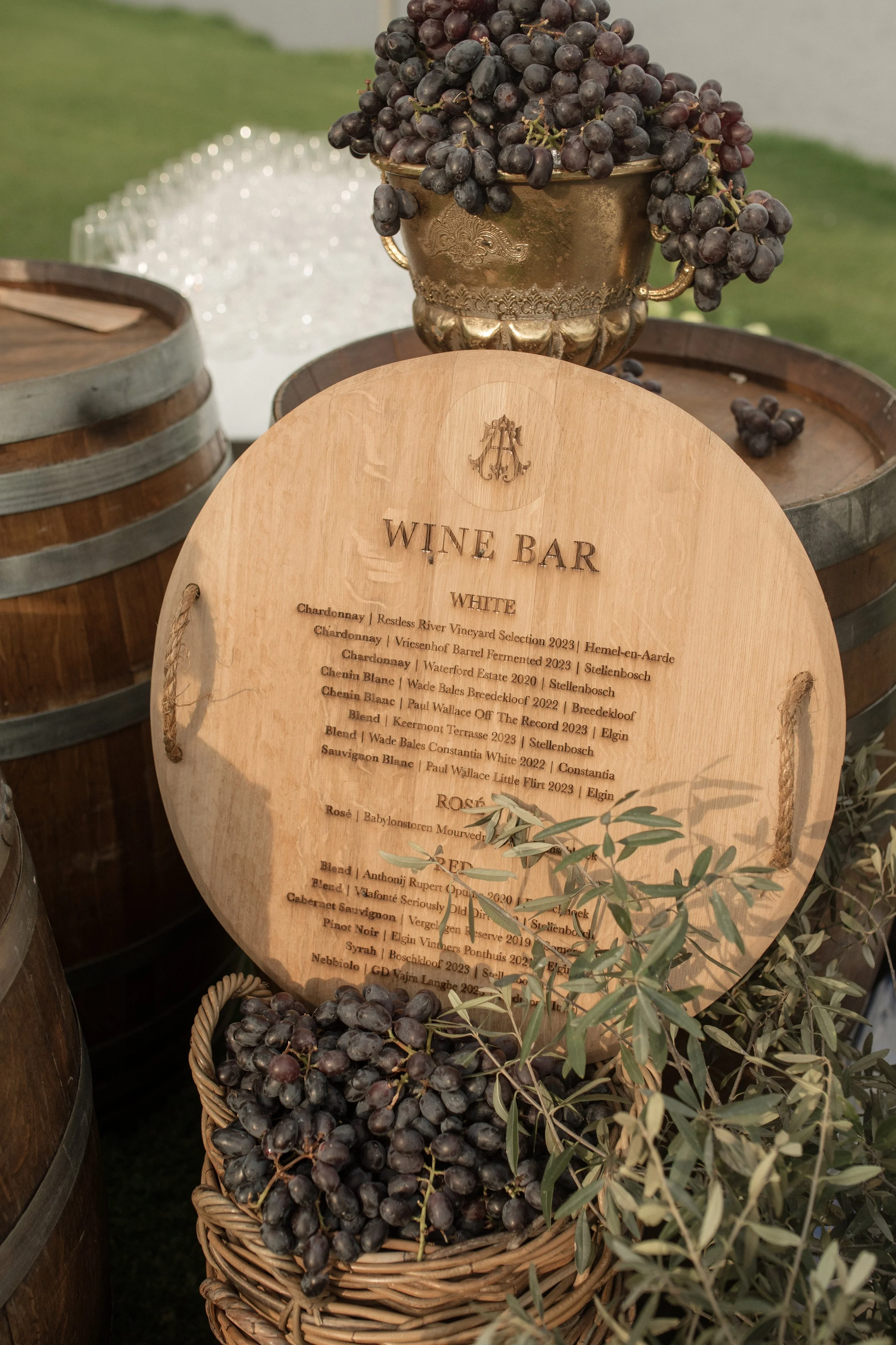 A basket of dark purple grapes with a background of wine barrels and a wooden sign listing various wine options, set outdoors with some leafy greenery.