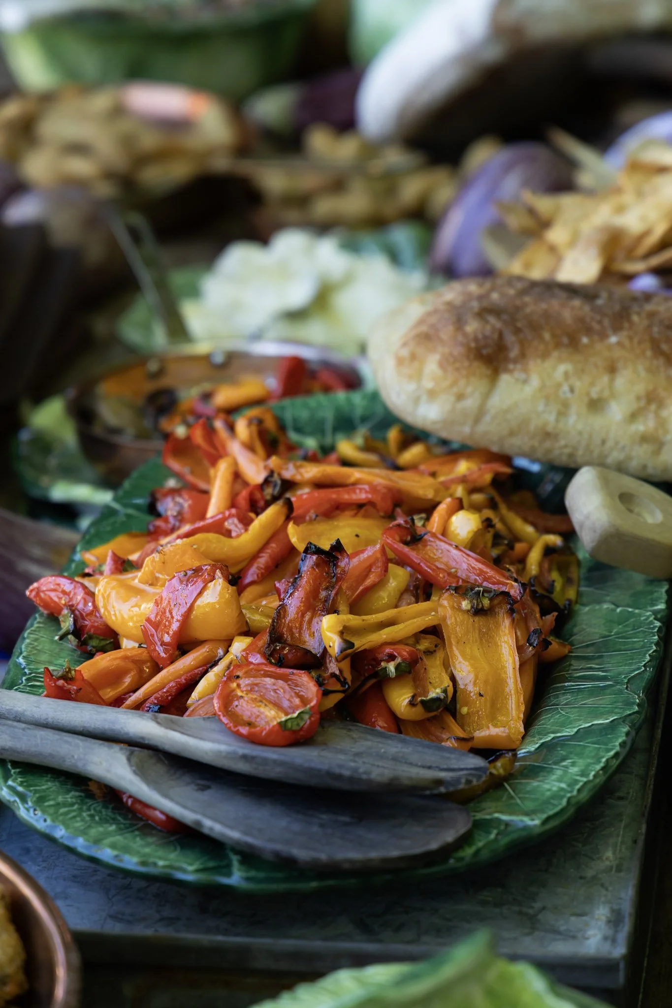 Colorful roasted peppers on a green leaf-lined plate with a bread roll and sides in the background.