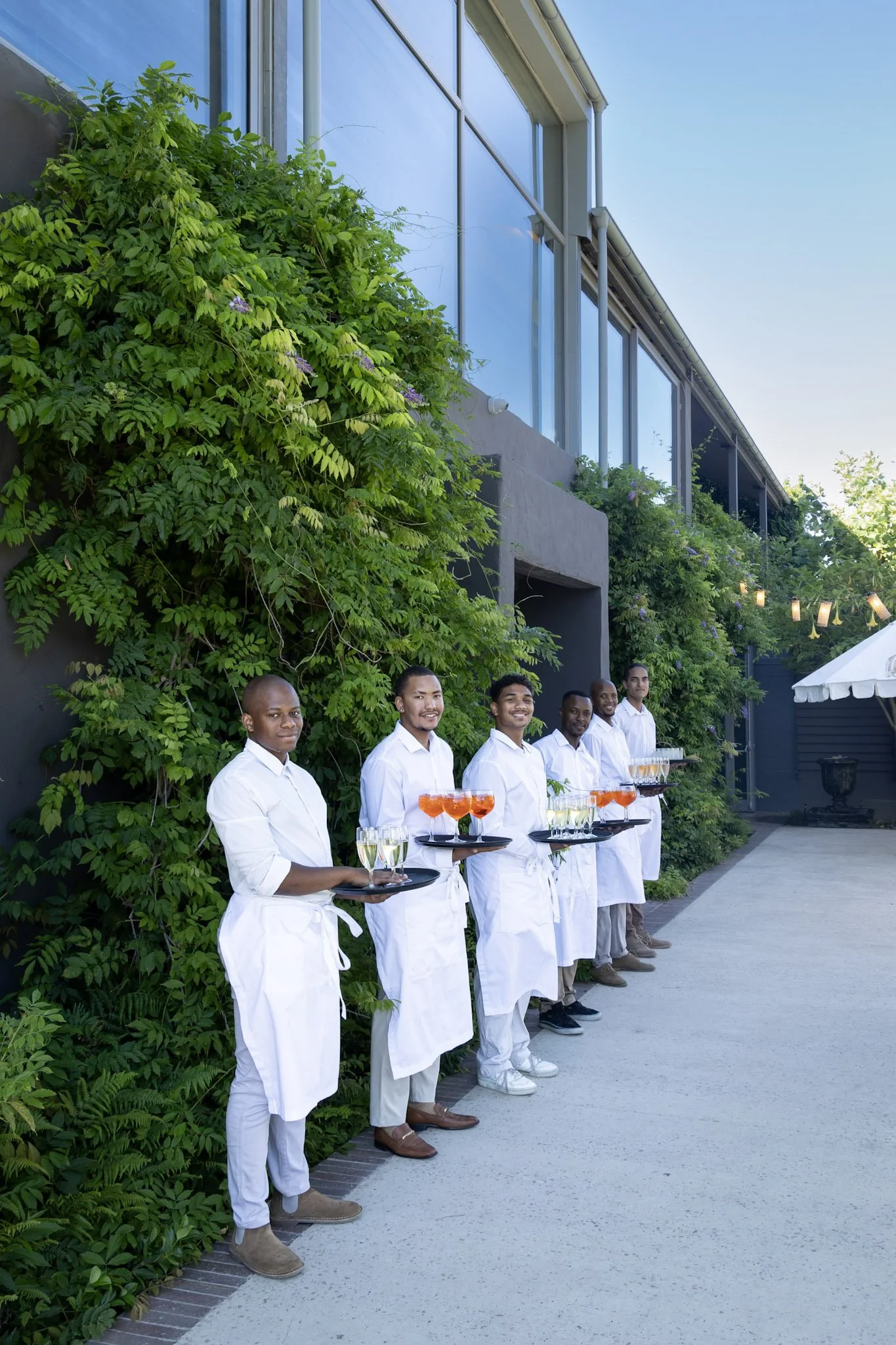 Five restaurant servers dressed in white shirts and aprons, holding trays with cocktails, standing outdoors in front of a building with green foliage on the wall.