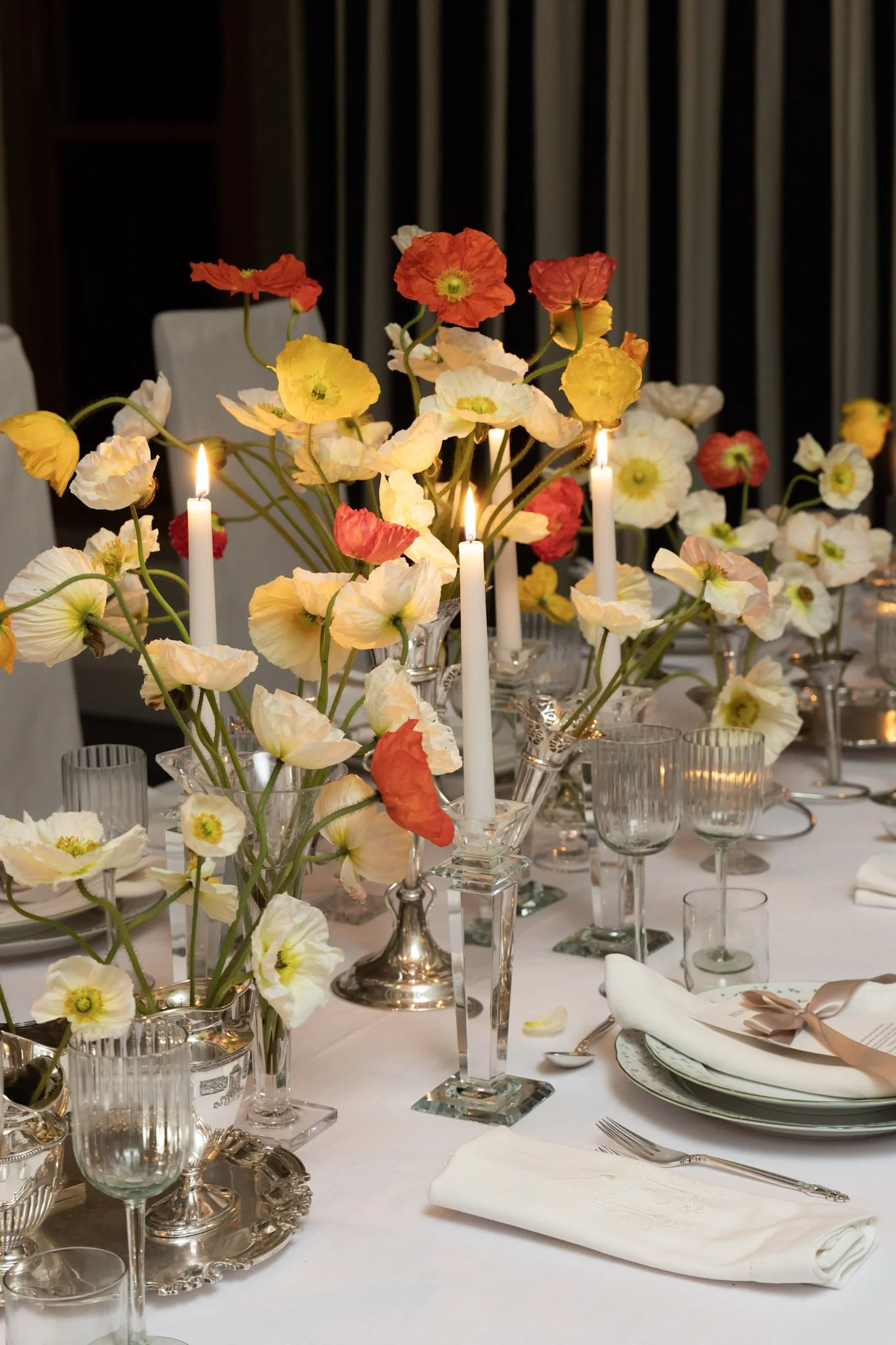 Elegant table setting with white tablecloth, floral centerpiece with colorful poppies, tall white candles, glassware, plates, and silverware.