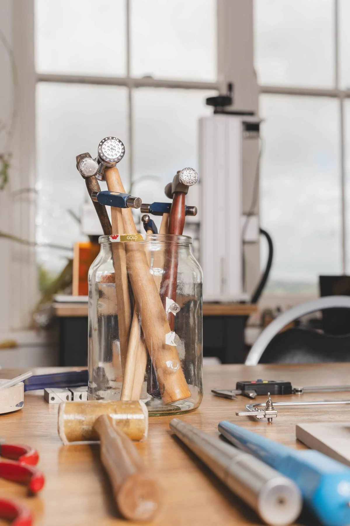 A glass jar filled with woodworking and carving tools on a workshop table, with additional tools and equipment in the background.