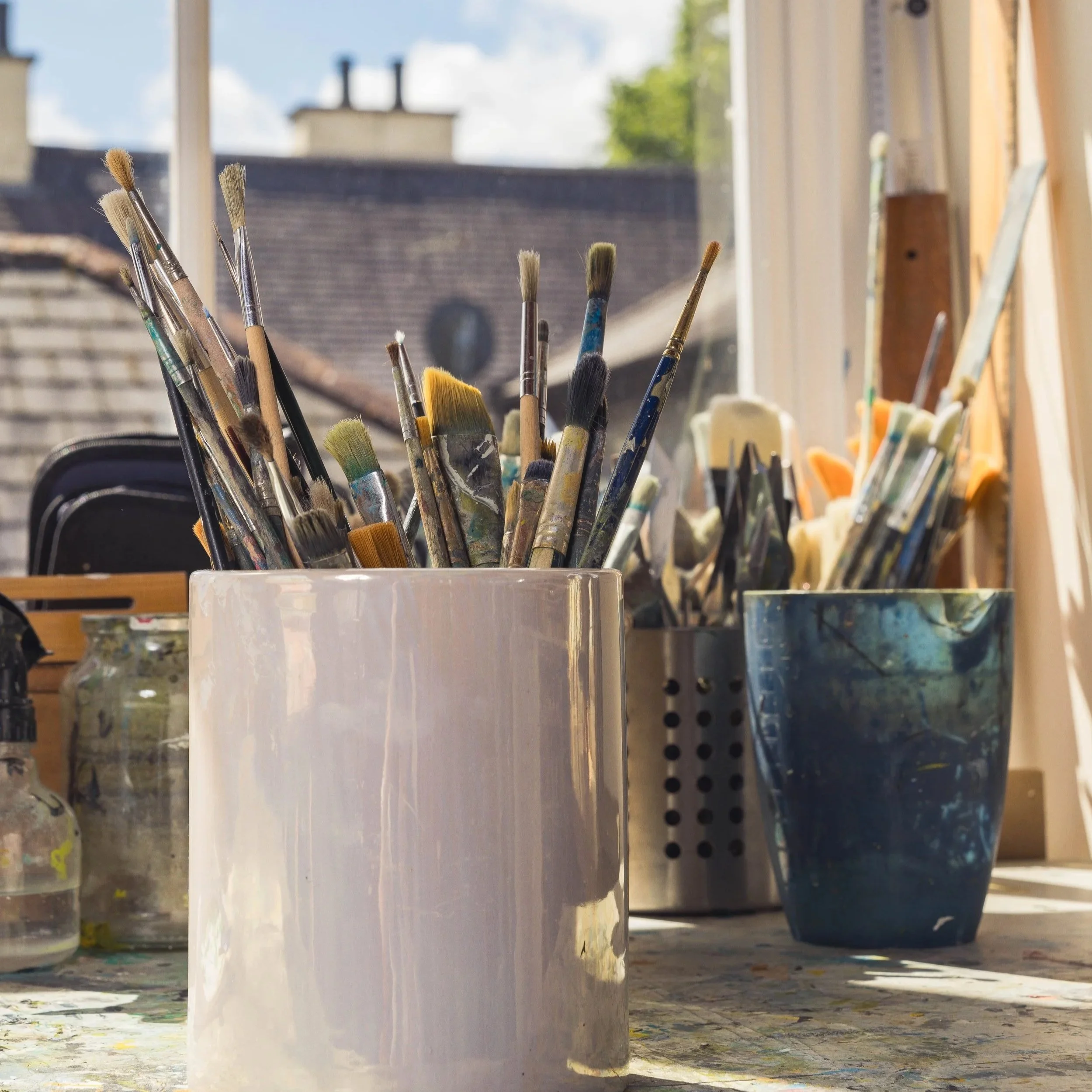 A collection of paintbrushes in a container on a work table with various jars of paint in the background and a window revealing a building and sky outside.