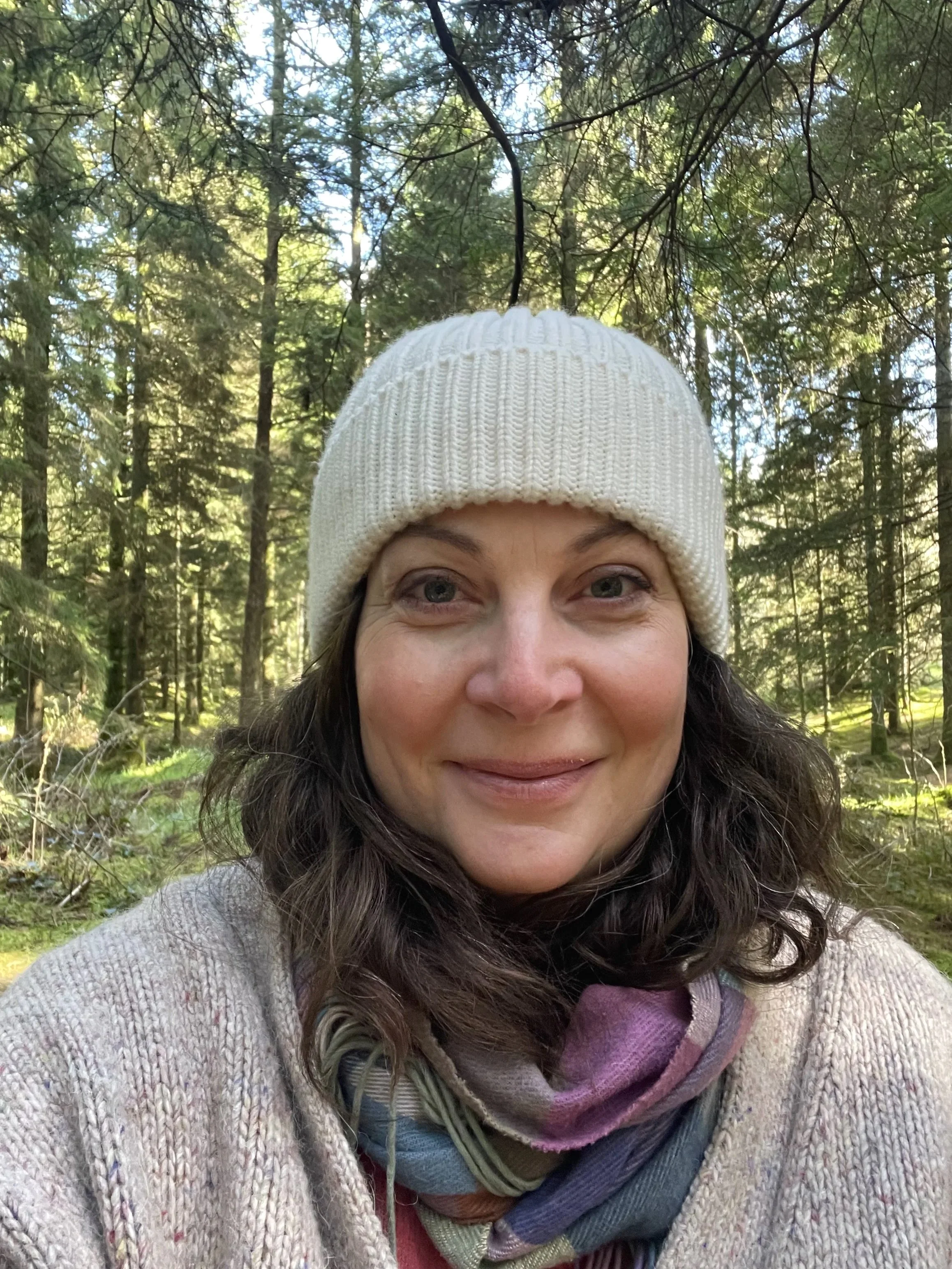 A woman with dark curly hair and light skin smiling outdoors in a forest, wearing a cream-colored knit hat, a cream sweater, and a colorful scarf.