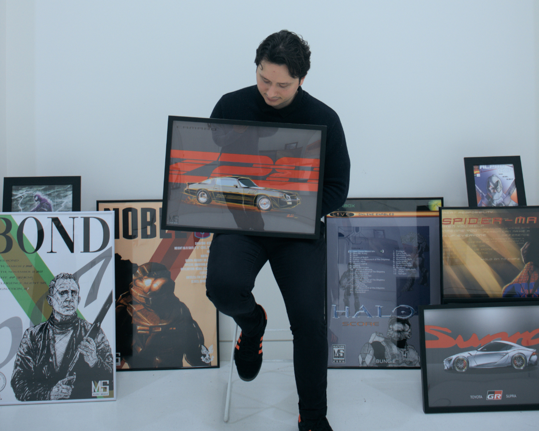 Person holding a framed artwork of a car, surrounded by other framed posters of video game and movie characters and vehicles in a white room.