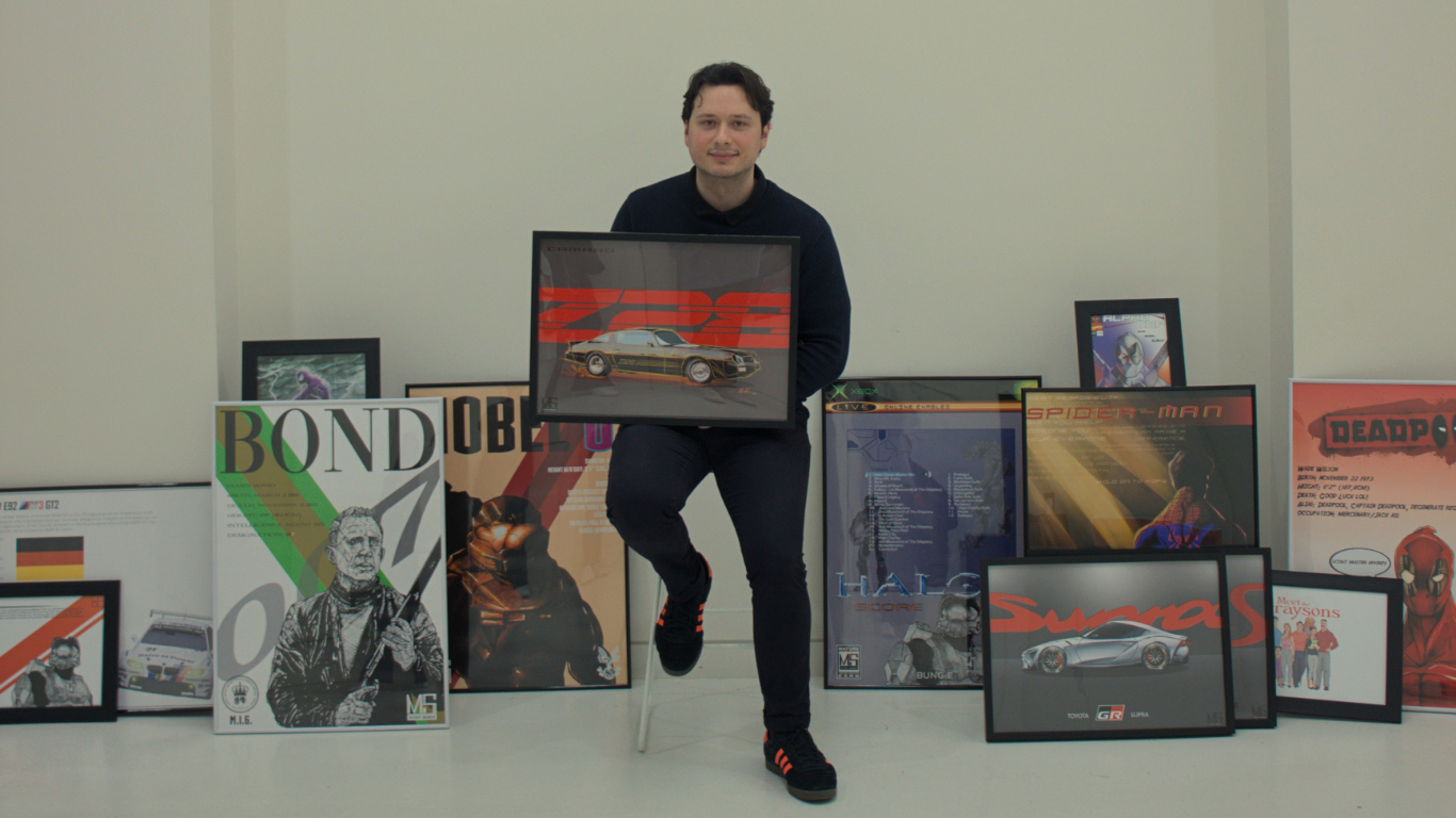 A man sitting on a white chair, holding a framed picture of a car, surrounded by framed posters of movies and video games displayed on the floor and wall behind him.