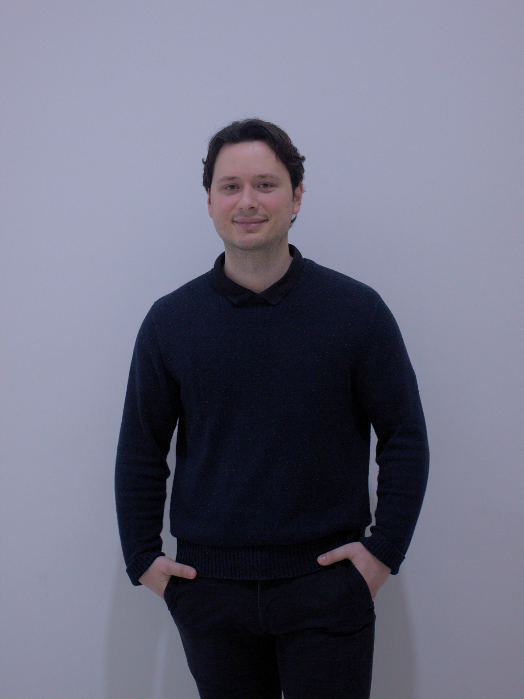 A young man with dark hair, wearing a black sweater and dark pants, standing in front of a plain light-colored wall, smiling slightly with hands in pockets.