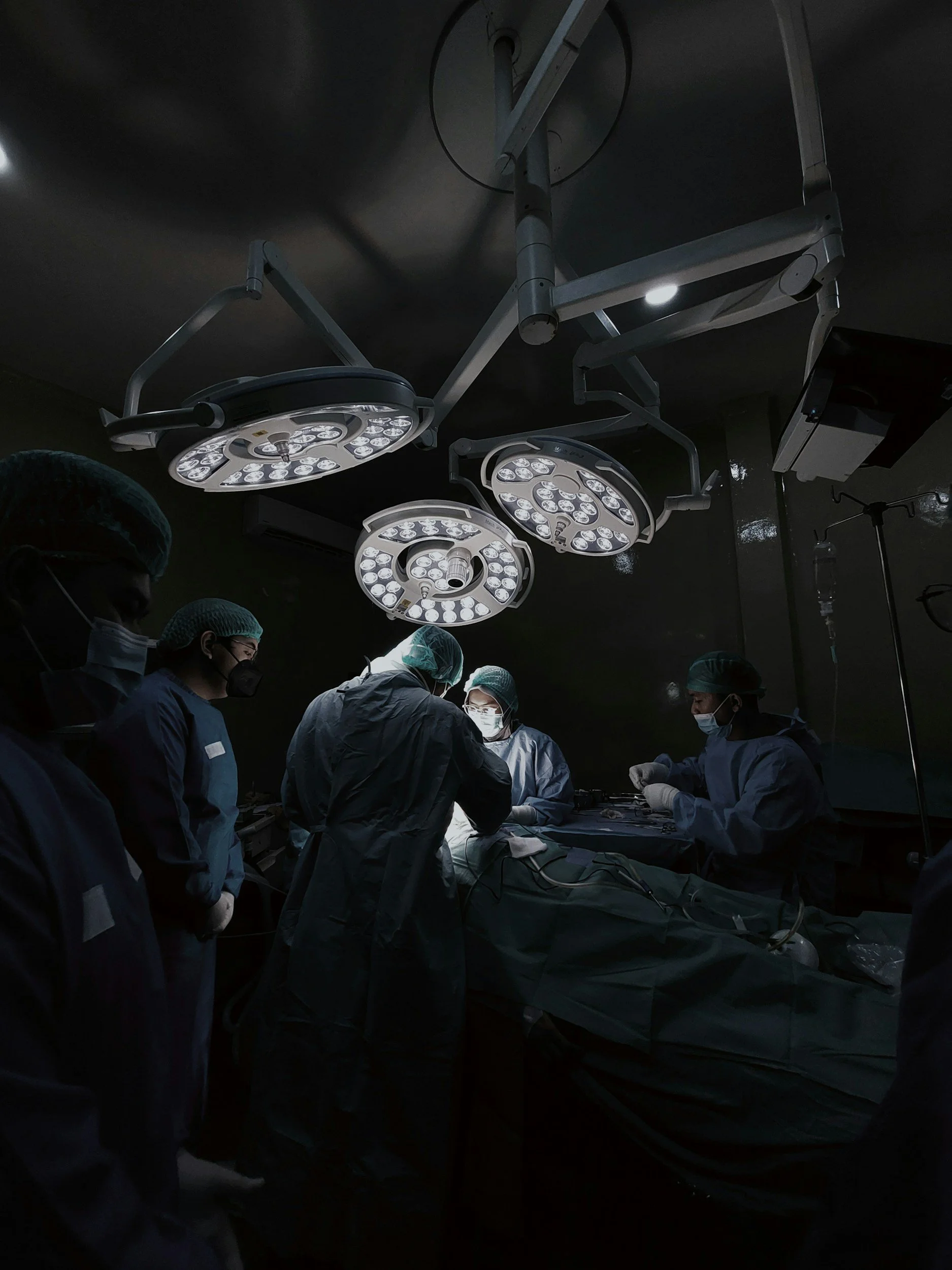 A surgical team performing an operation in an operating room with overhead surgical lights and medical staff wearing surgical scrubs and masks.