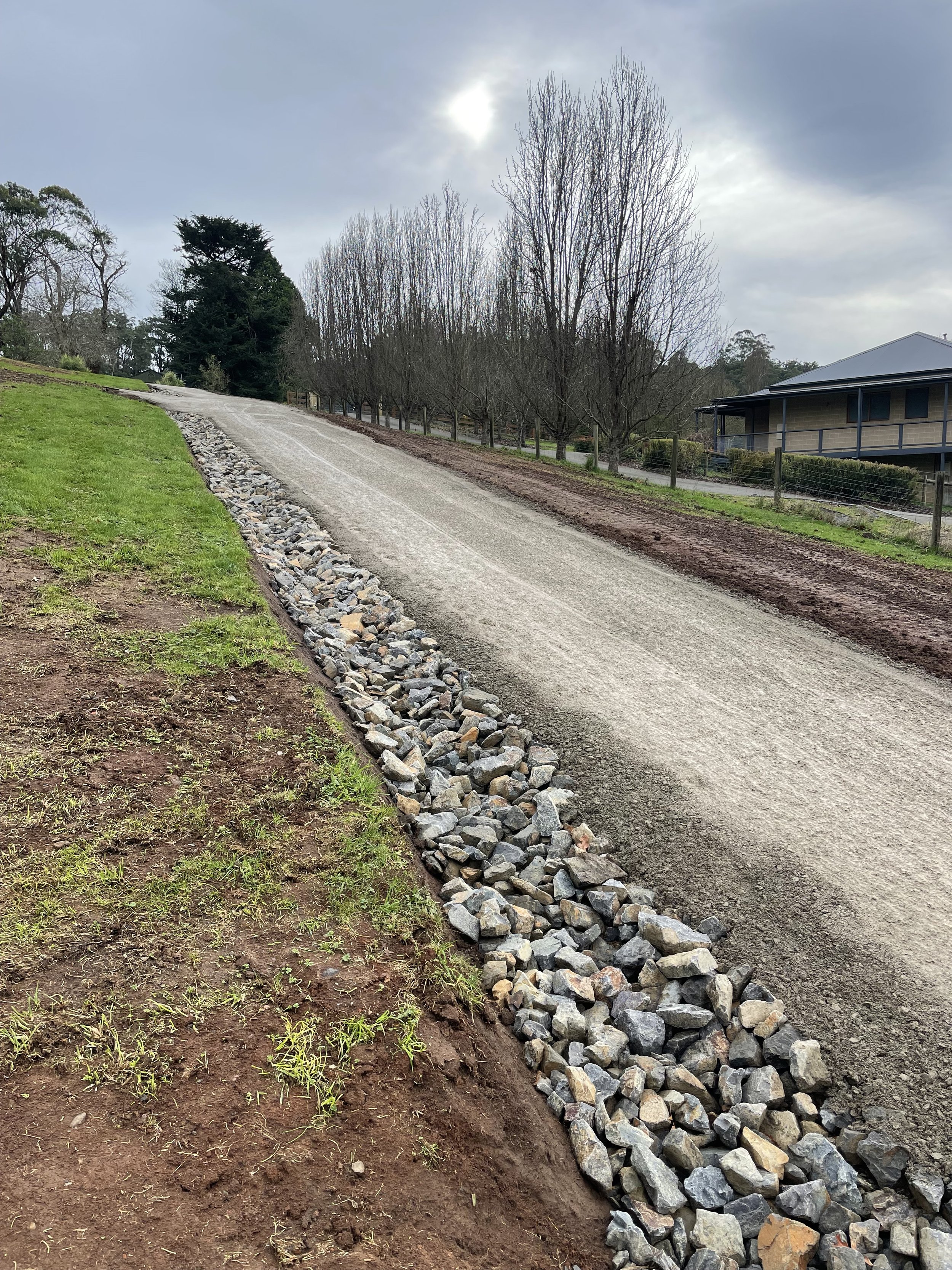 A gravel driveway on a slope with a strip of rocks on one side, lined with leafless trees and a house with a porch, under a cloudy sky with the sun faintly visible.