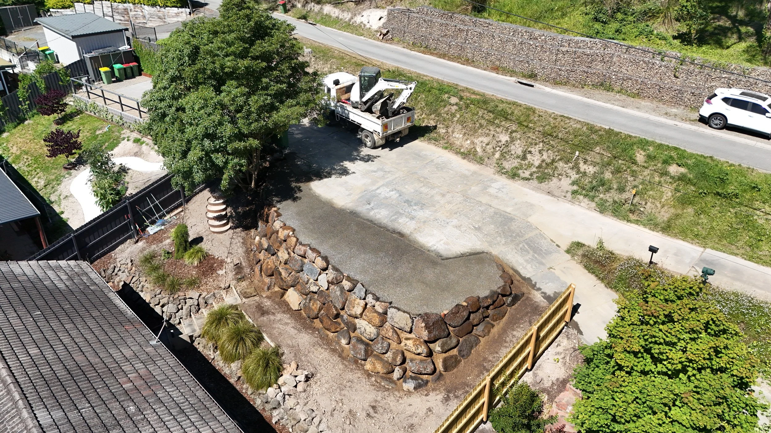 A backyard with a tree, a stone border, a lawn, and a fence. There is a small construction area with dirt and rocks, a truck with a mini excavator, and parked cars along the street.