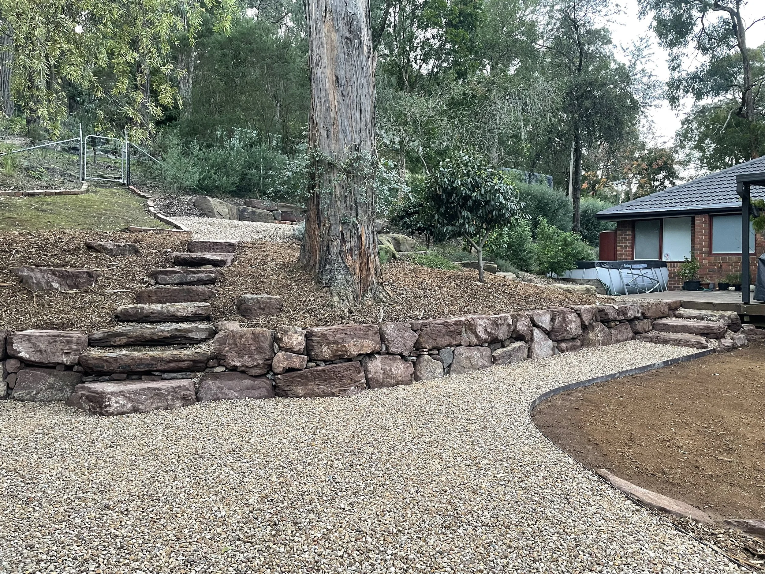 A backyard garden area with stone steps, a large tree, shrubs, and a house with a tiled roof and sliding glass door.