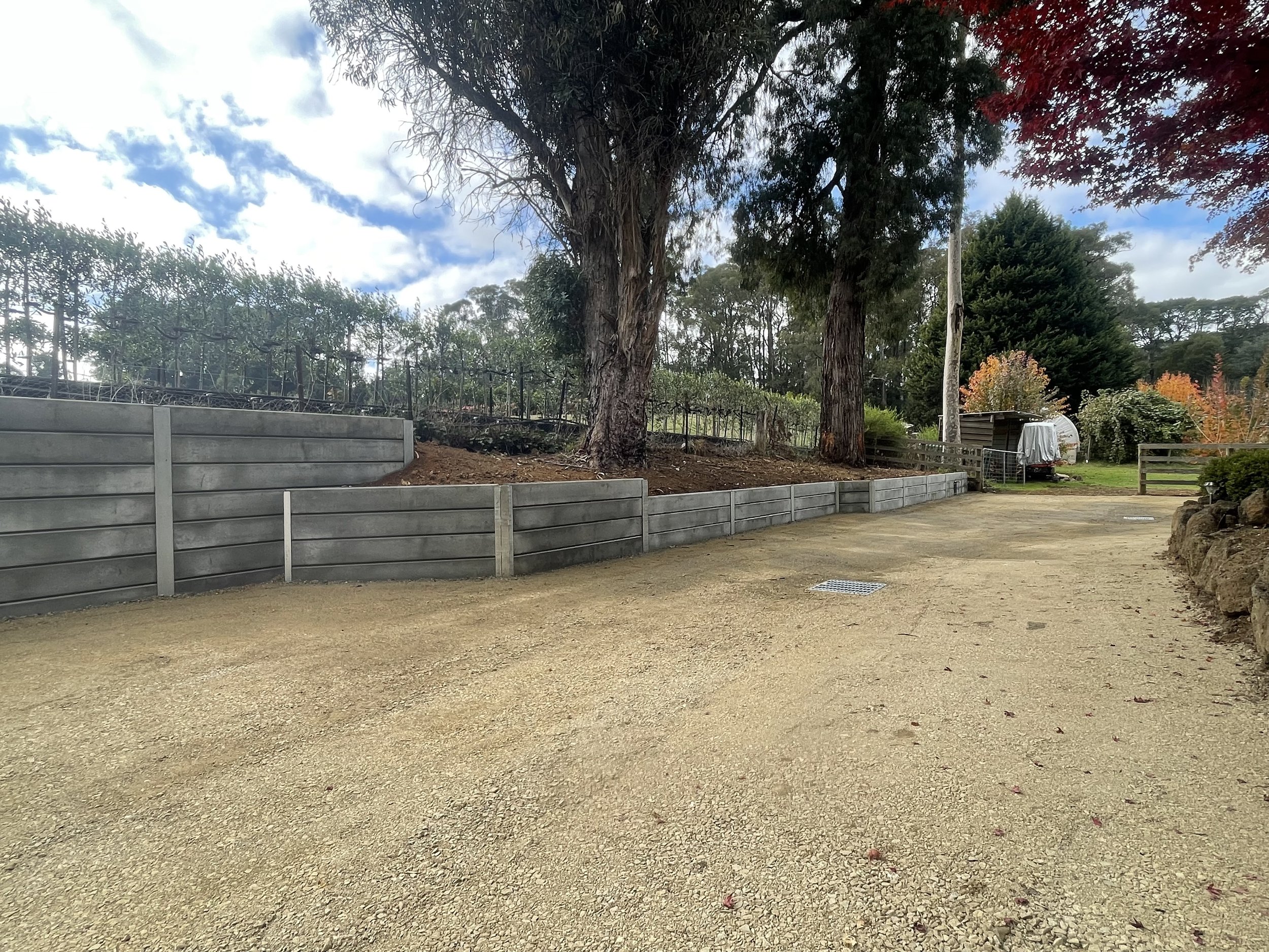 A dirt path surrounded by trees with green, red, and yellow foliage. Behind the trees, there is a house or shed and a white trailer. The sky is partly cloudy with patches of blue.