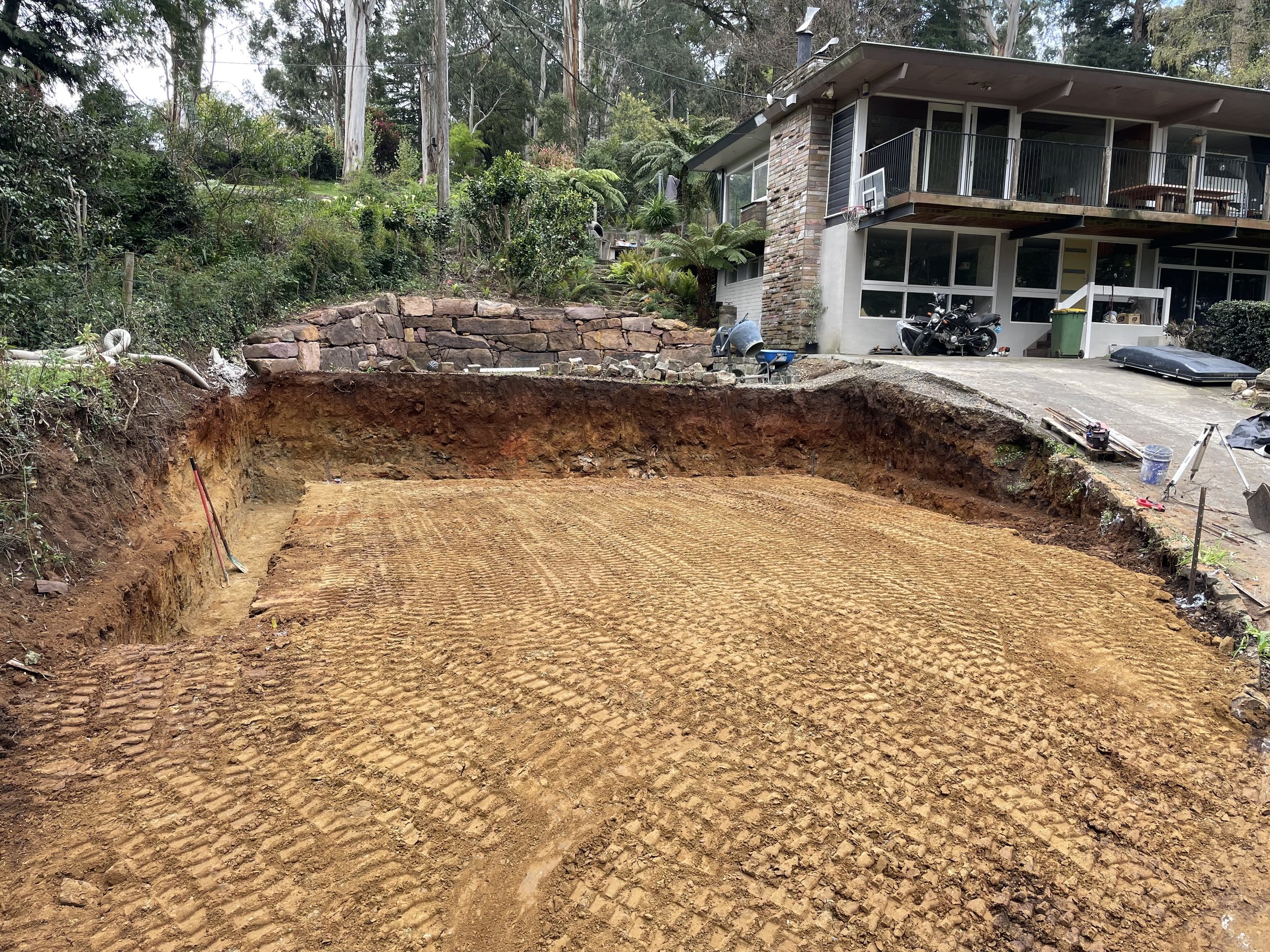 A residential backyard undergoing landscape or construction work, featuring a large excavated area with exposed dirt and tire tracks, a modern house with a balcony, parked motorcycle, tools, and construction materials.