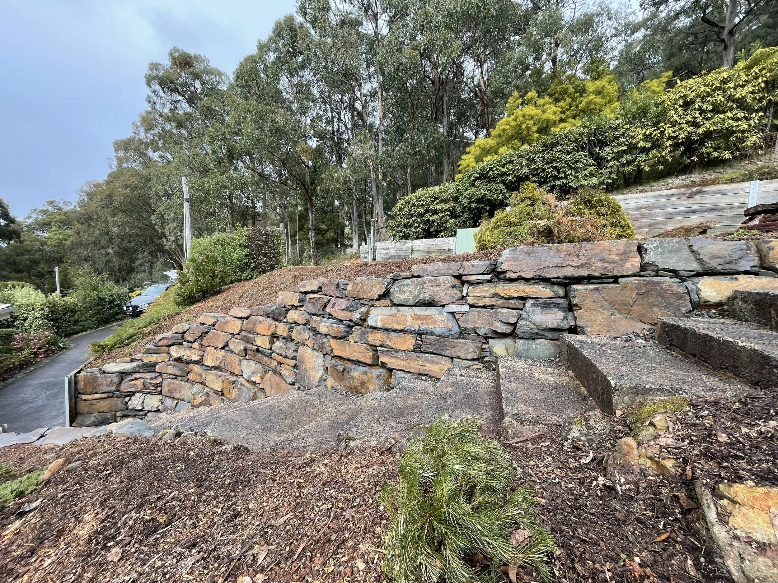 A stone retaining wall with a garden and trees behind it, in a residential area, during daylight.