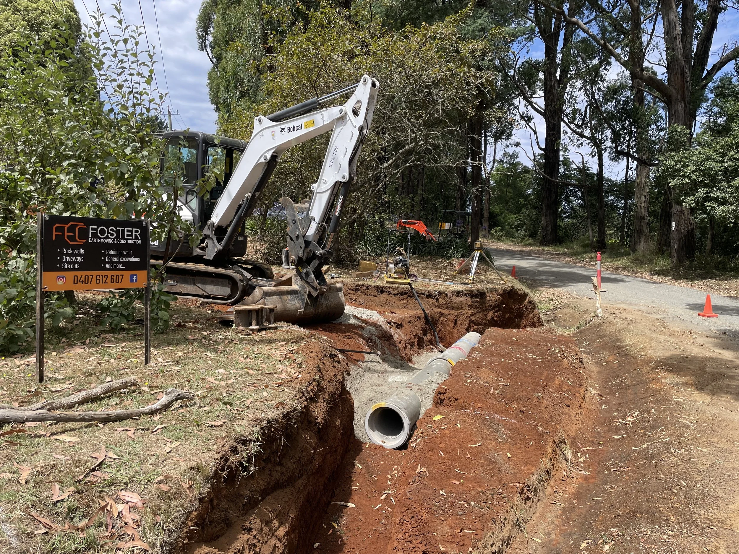 Construction site with a mini excavator digging a trench through a residential street lined with trees. Construction equipment, safety cones, and a sign for earthmoving and construction services are visible.