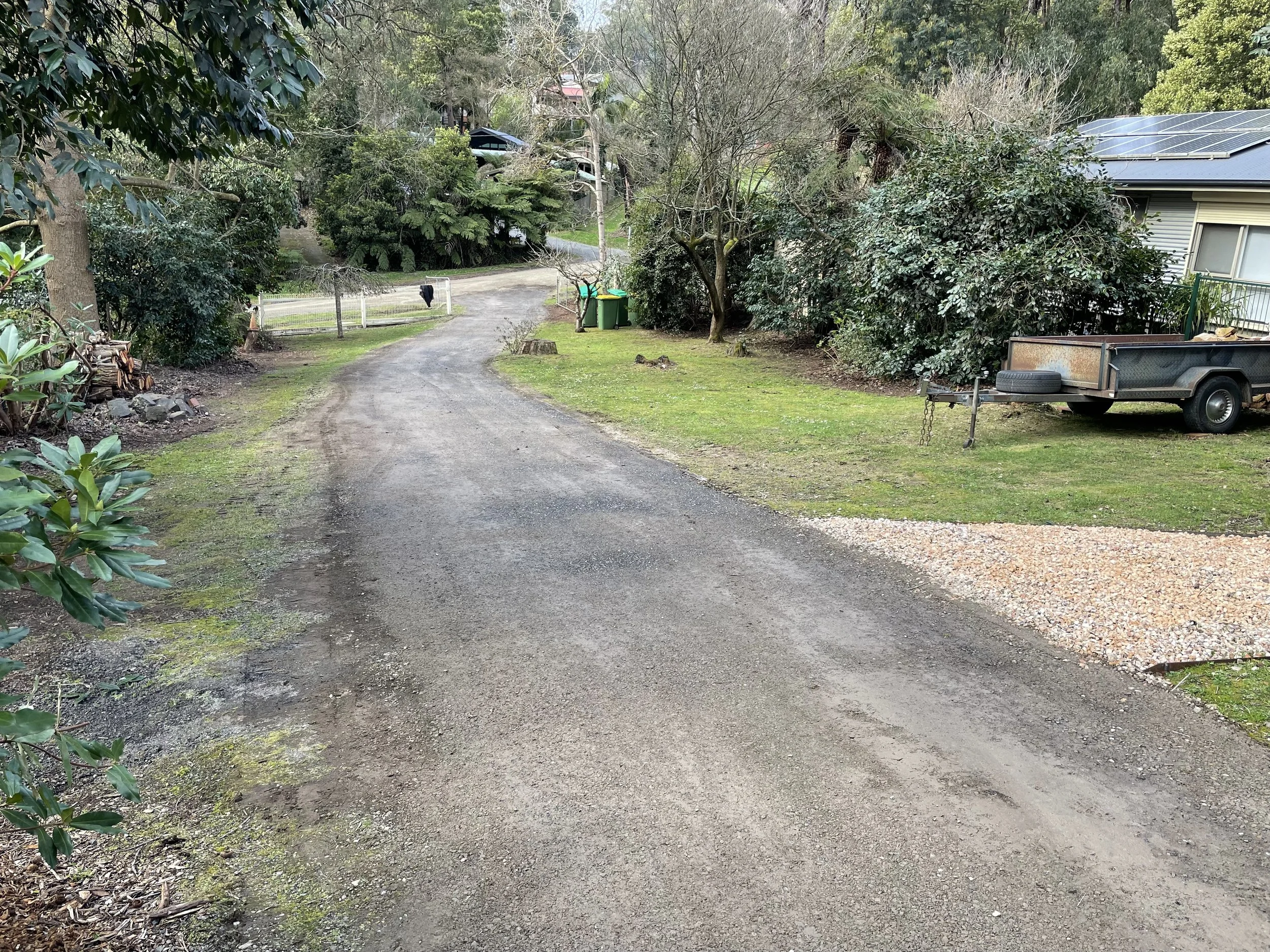 A gravel driveway winding through a wooded backyard with trees and shrubs, a small trailer parked on the right, and green bushes near a house with a metal roof in the background.