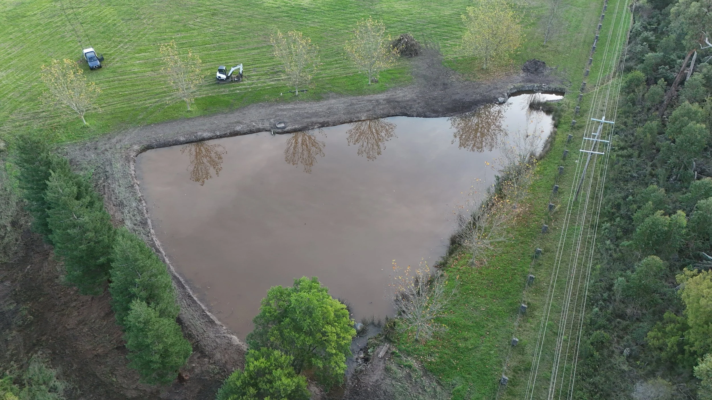 Aerial view of a construction site next to a pond, with trees, grass, and power lines nearby. A small excavator and a pickup truck are present on the grassy area.