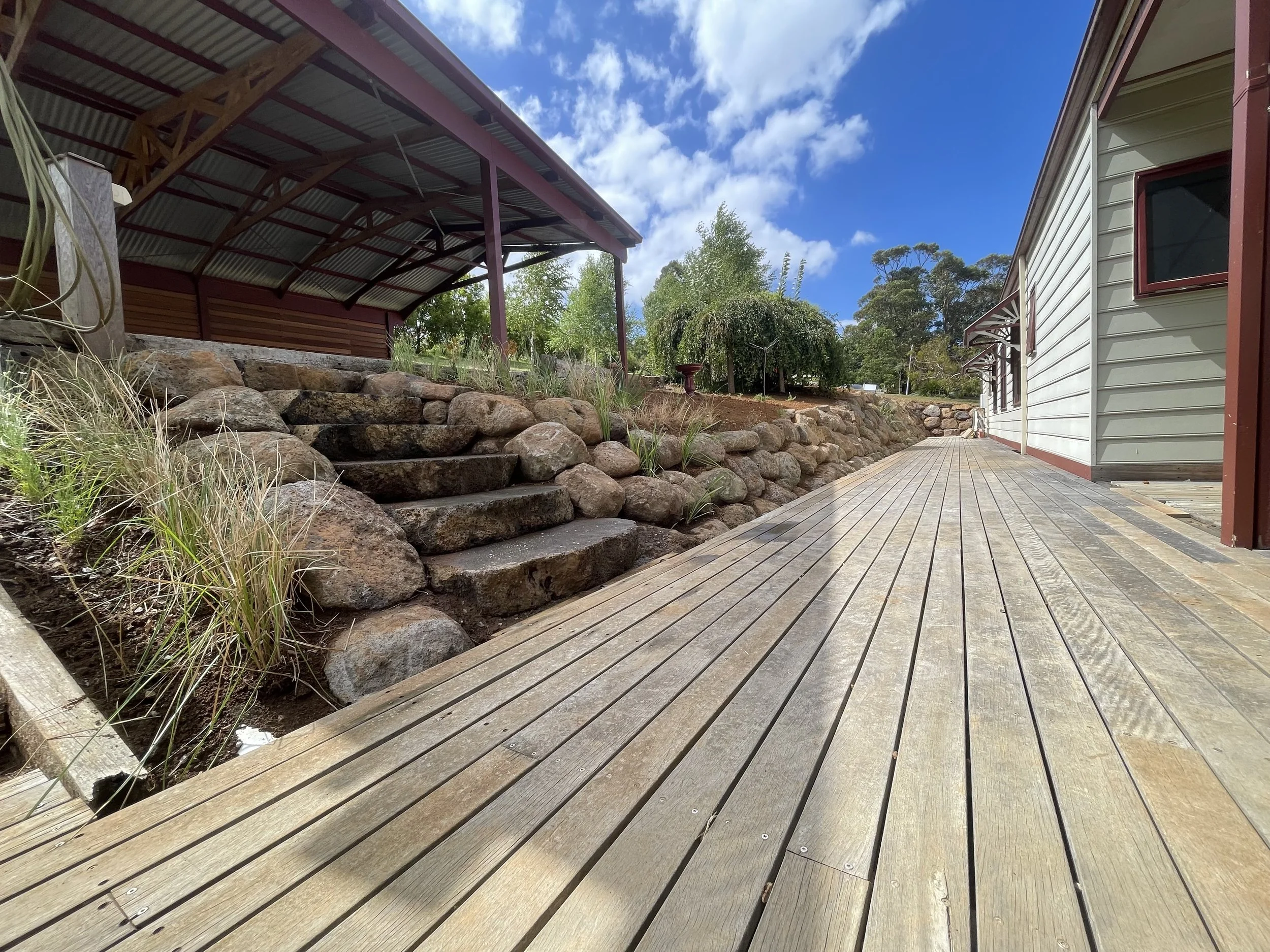 Backyard with a wooden deck, stone steps, a rock wall, a grassy area with trees, and a house with white siding and red trim under a partly cloudy sky.