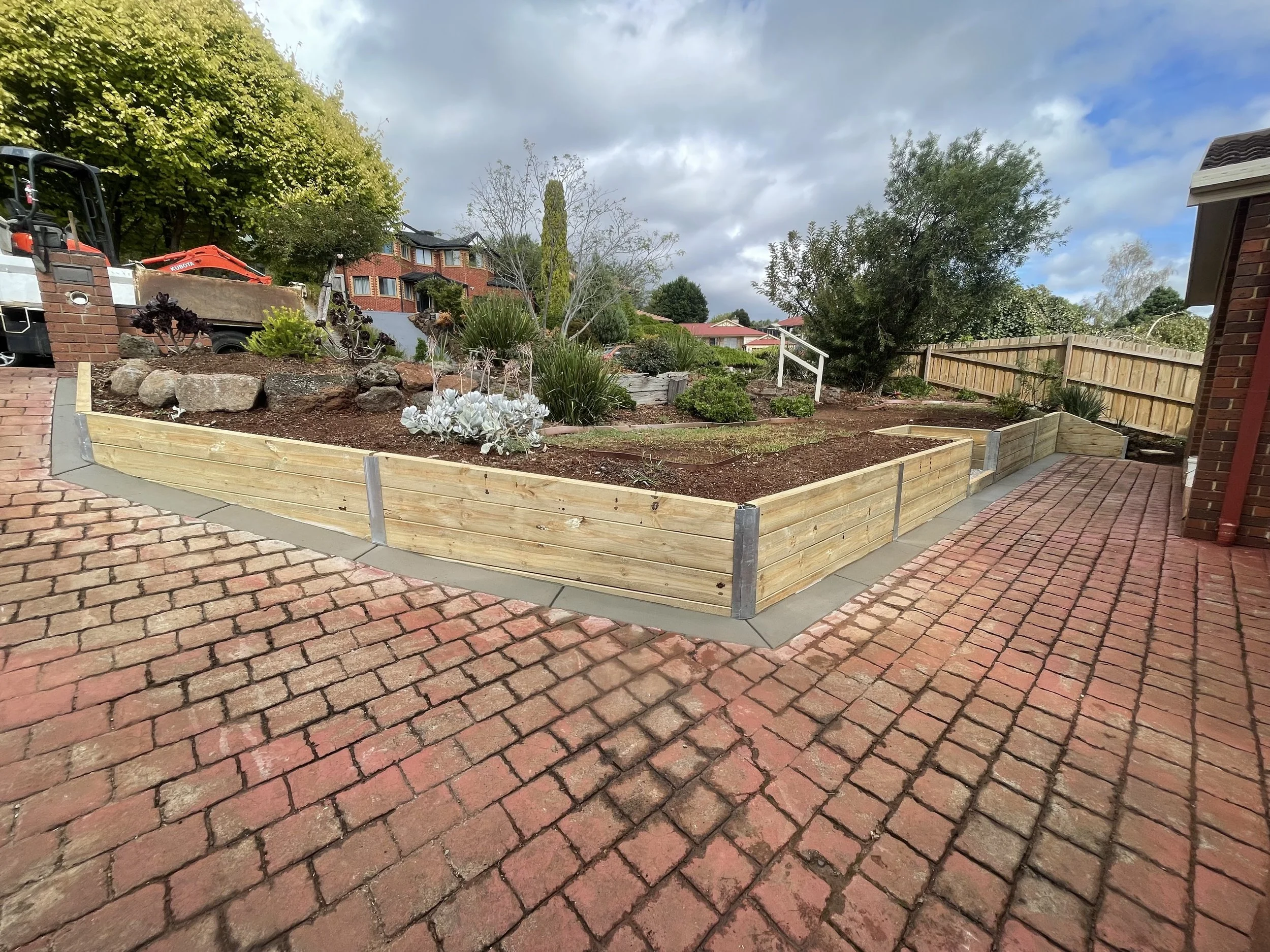 A backyard garden with raised beds, plants, trees, and a brick fence surrounding the area, with houses and a cloudy sky in the background.