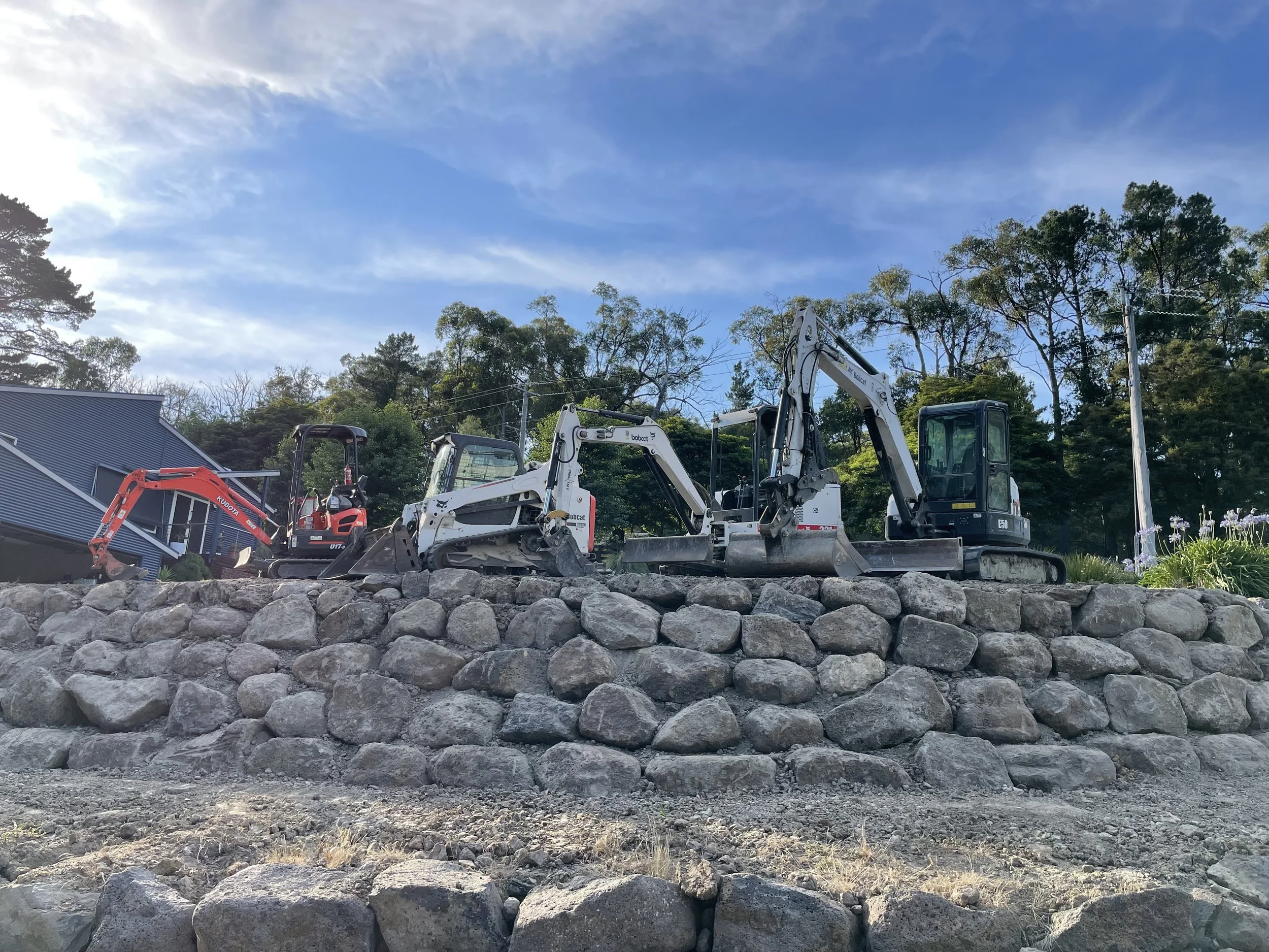 Construction site with several mini excavators and large rocks in the foreground, trees and utility poles in the background under a partly cloudy sky.