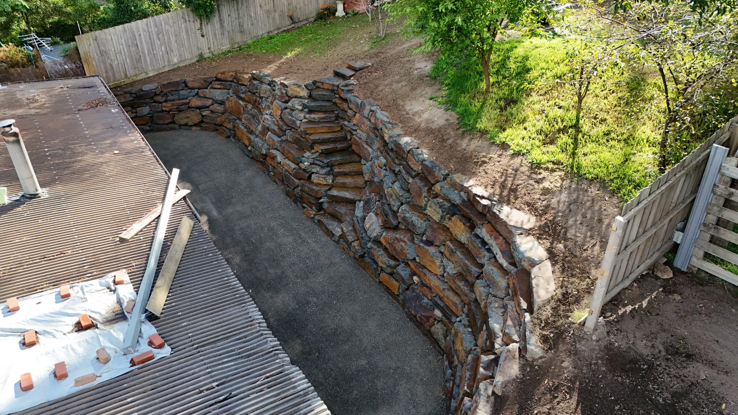 A stone retaining wall under construction in a backyard. The wall is made of variously sized flat stones and separates a gravel area from a sloped area with grass and trees.