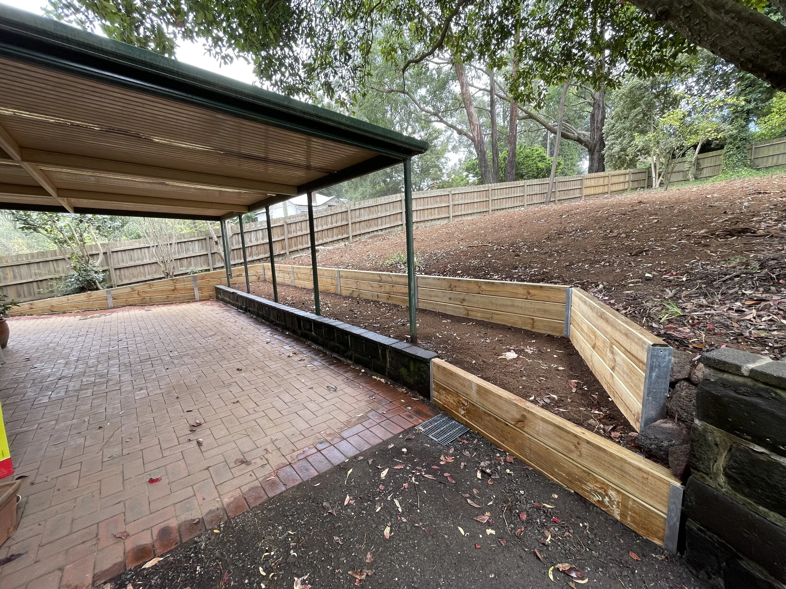 A backyard with a brick patio, a covered area with a metal roof, and a sloped yard enclosed by a wooden fence. There are trees and bushes in the background.
