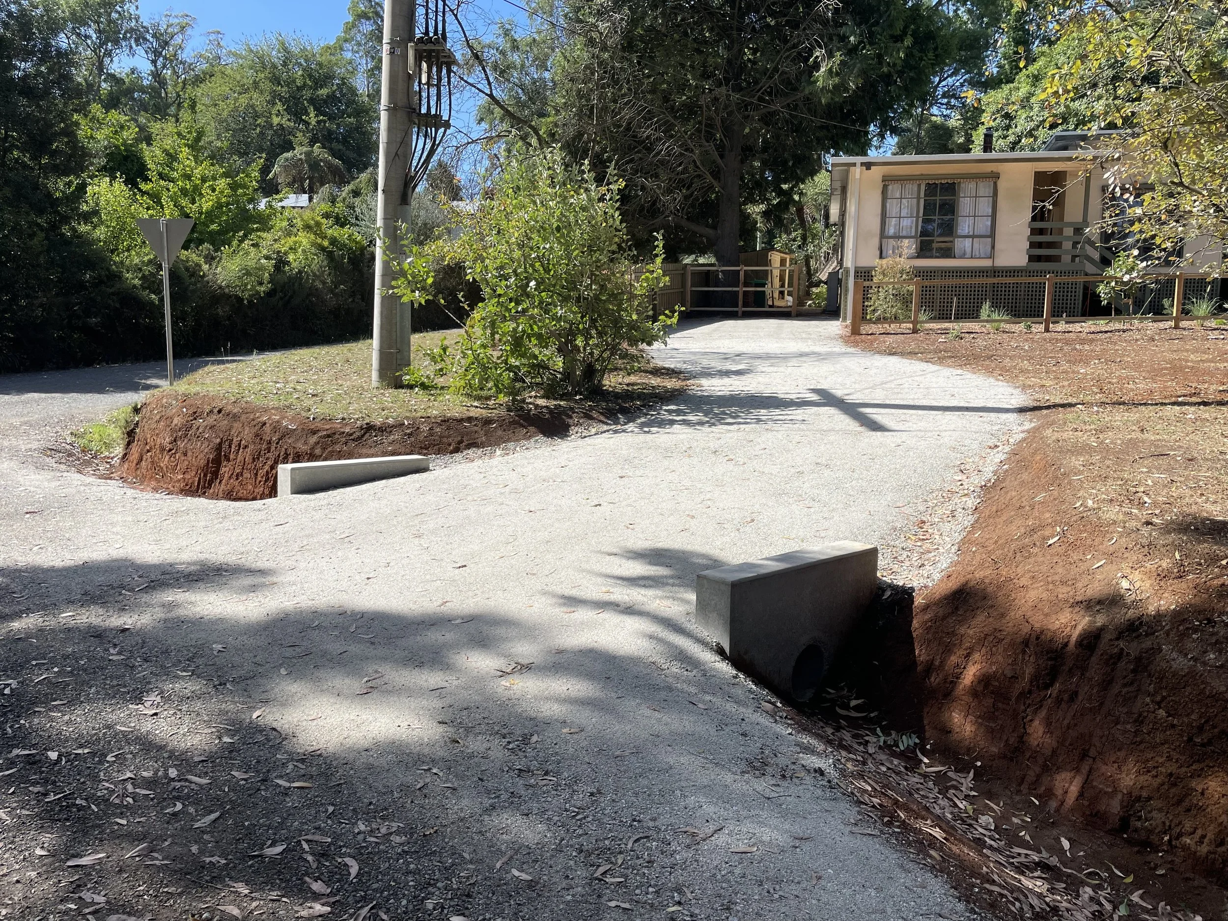 A dirt driveway with construction curb blocks, a utility pole, a wooden fence, trees, and a house with large windows and stairs outside.