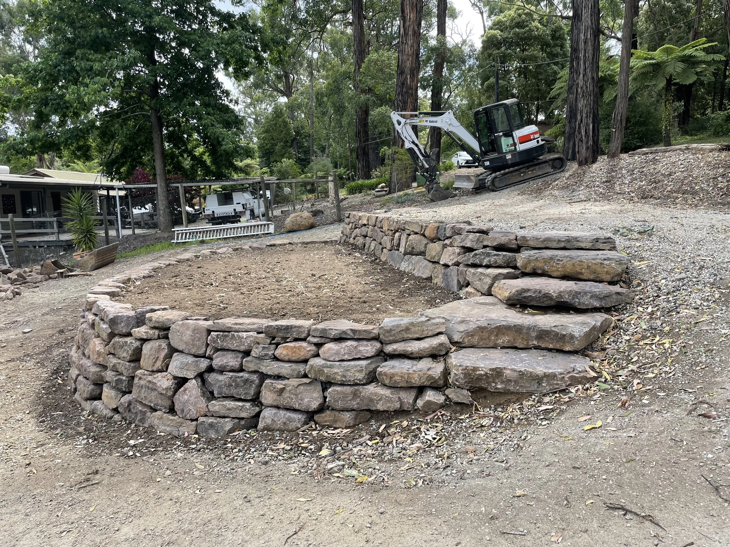 A construction site with a small excavator on a dirt area next to a large pile of rocks, surrounded by trees and a residential area with houses and cars in the background.