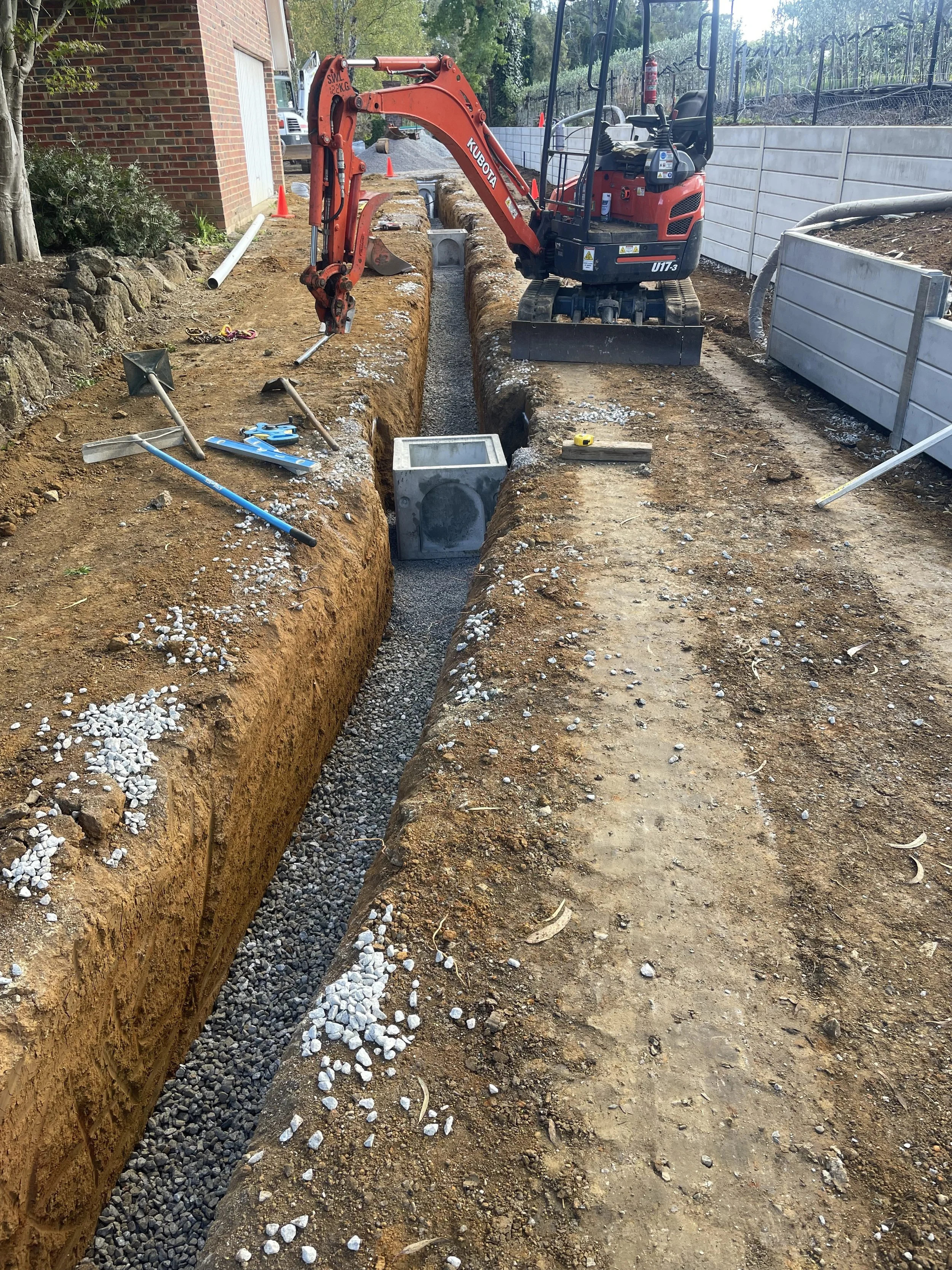 Construction site with a small excavator digging a trench for underground pipes, surrounded by dirt and construction tools, with safety cones and barriers.