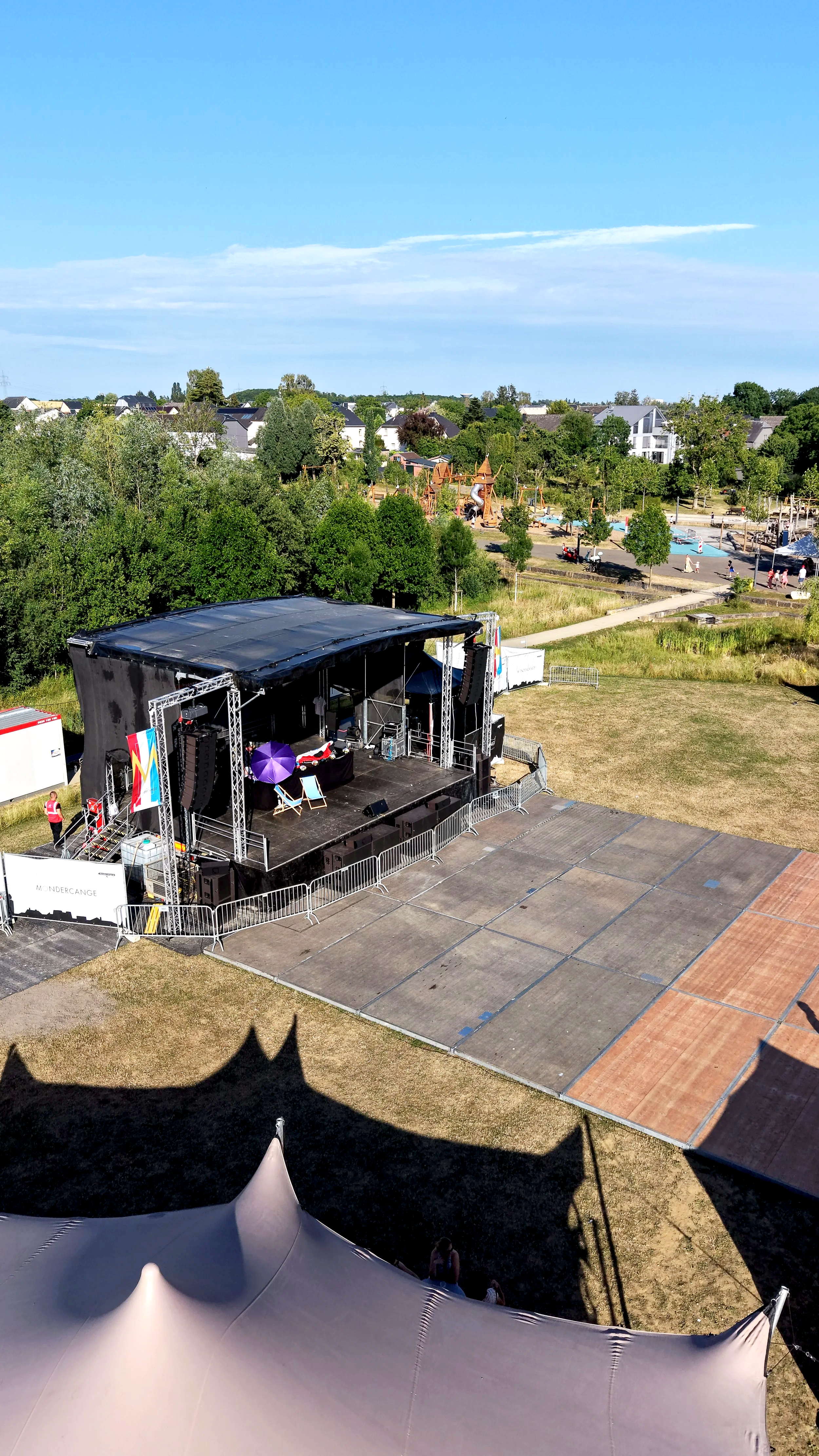 An outdoor event stage with sound equipment and a purple umbrella on a sunny day. There is a tent in the foreground and a grassy area with a playground and people in the background.