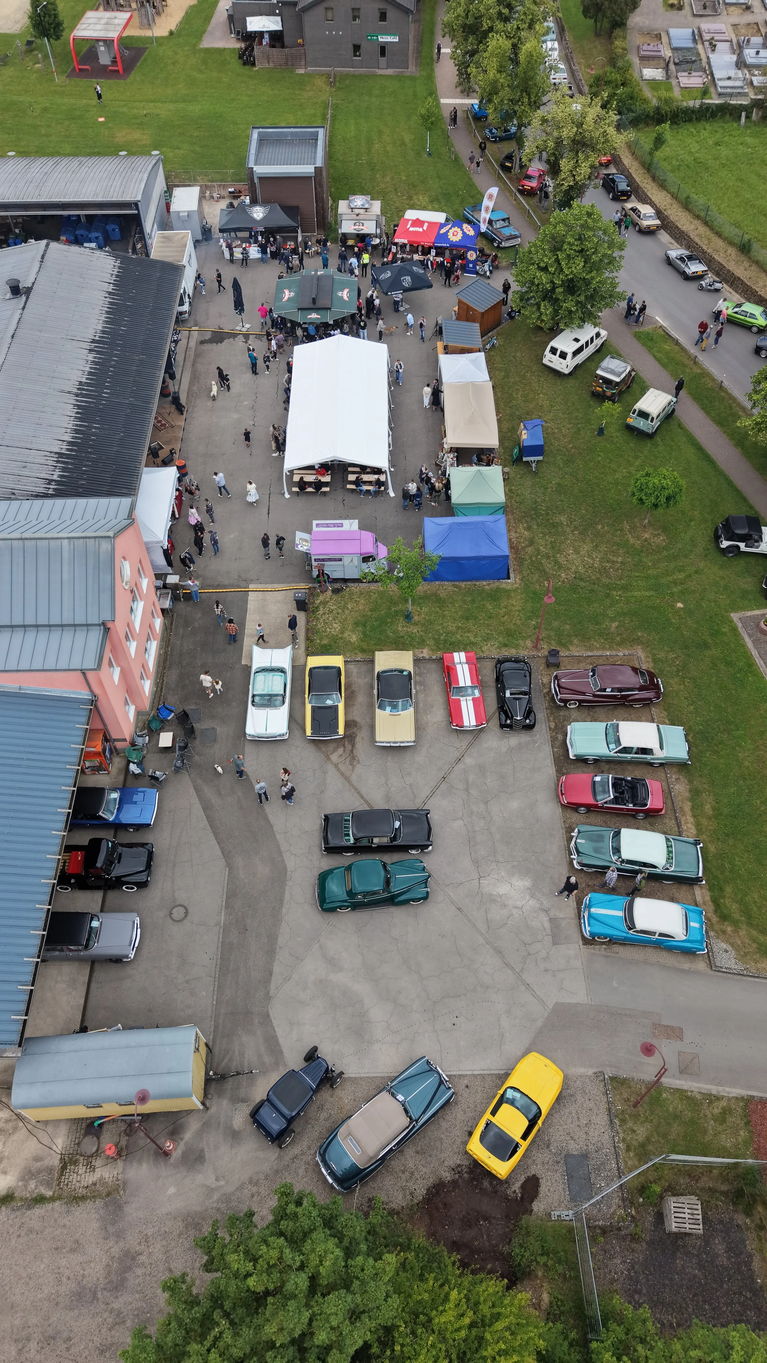 An aerial view of a car show or outdoor event with various tents, booths, and a crowd of people gathering in the center. There are several parked classic and vintage cars nearby, and some grassy areas with trees and pathways.