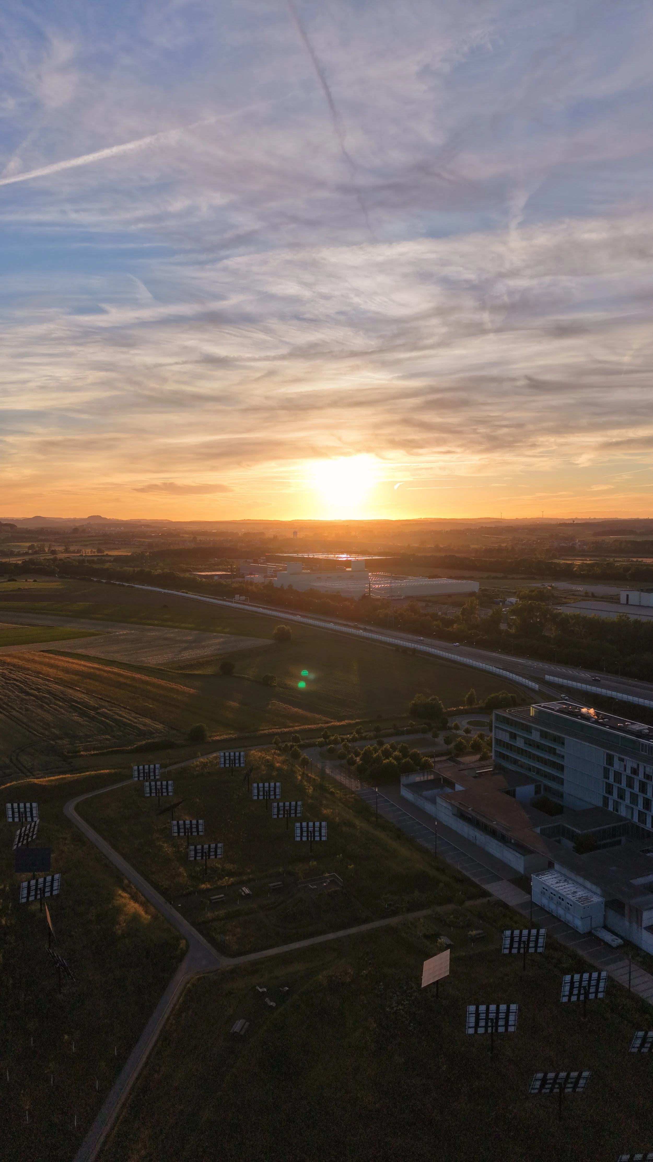 Sunset over a landscape with fields, roads, and buildings, with a sky filled with clouds and contrails.